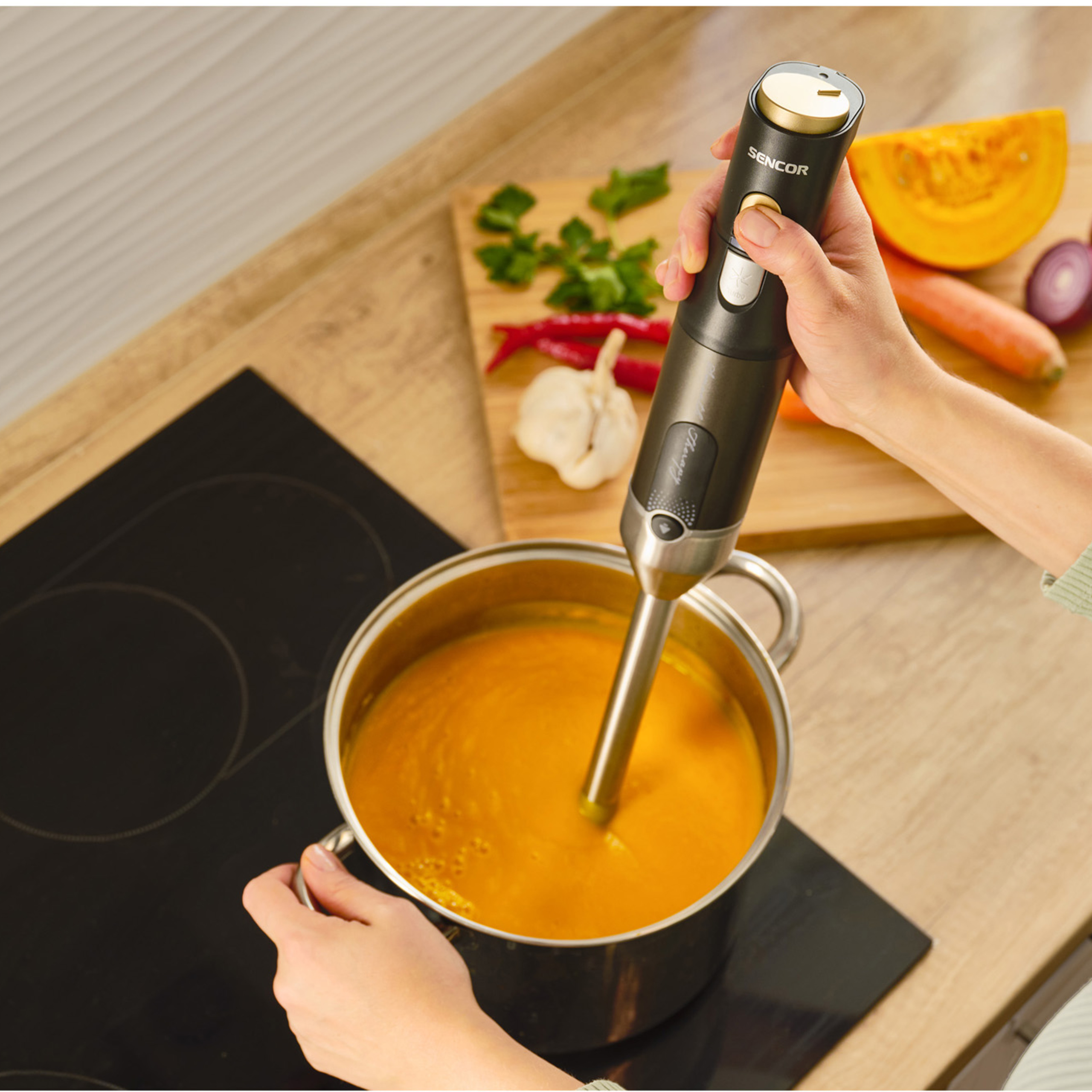 Eine Hand bedient einen Sencor-Stabmixer in einem Topf mit oranger Suppe auf einem Herd, im Hintergrund Gemüse.