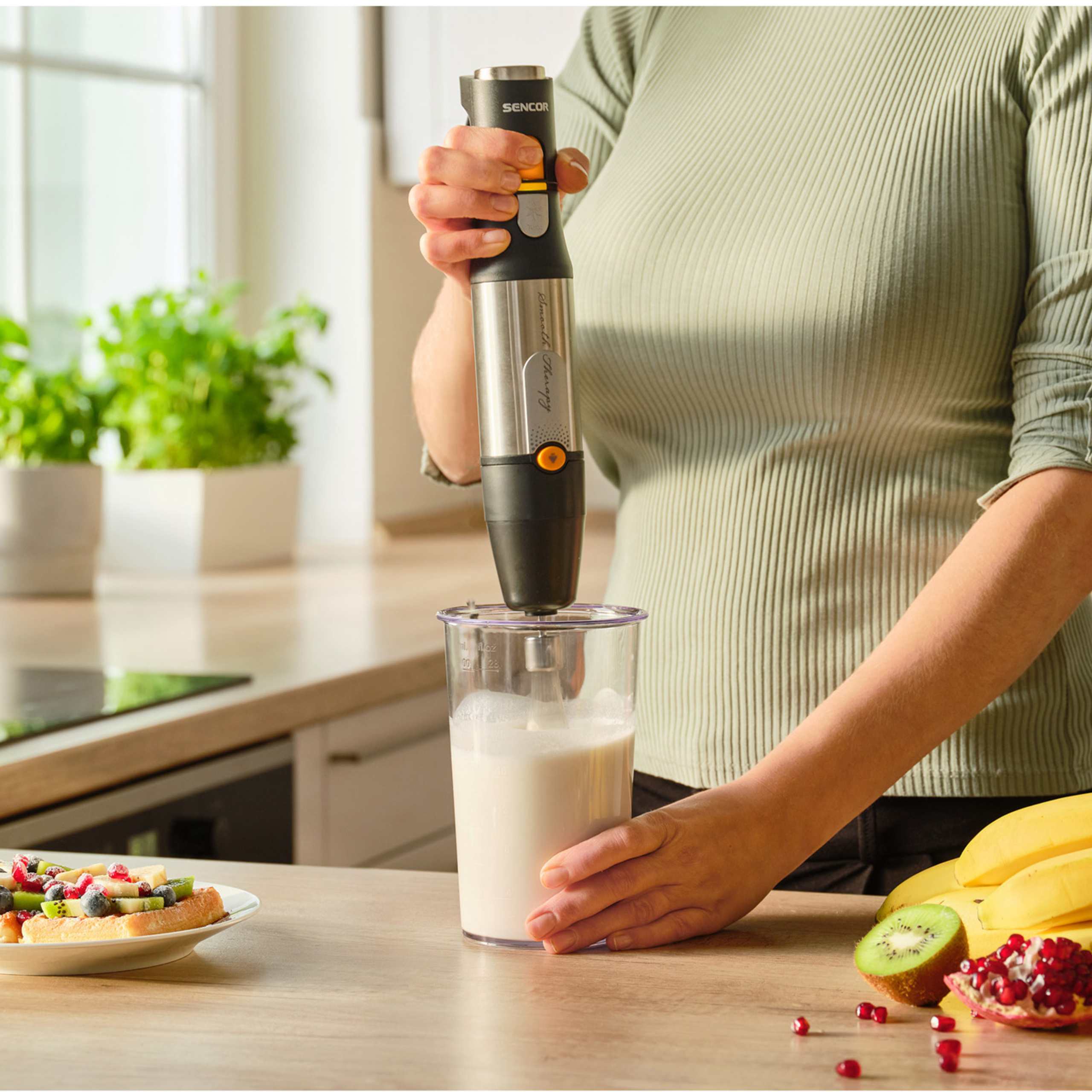 Una persona licúa una bebida blanca en un vaso alto con una batidora de mano, con fruta y un gofre cerca.