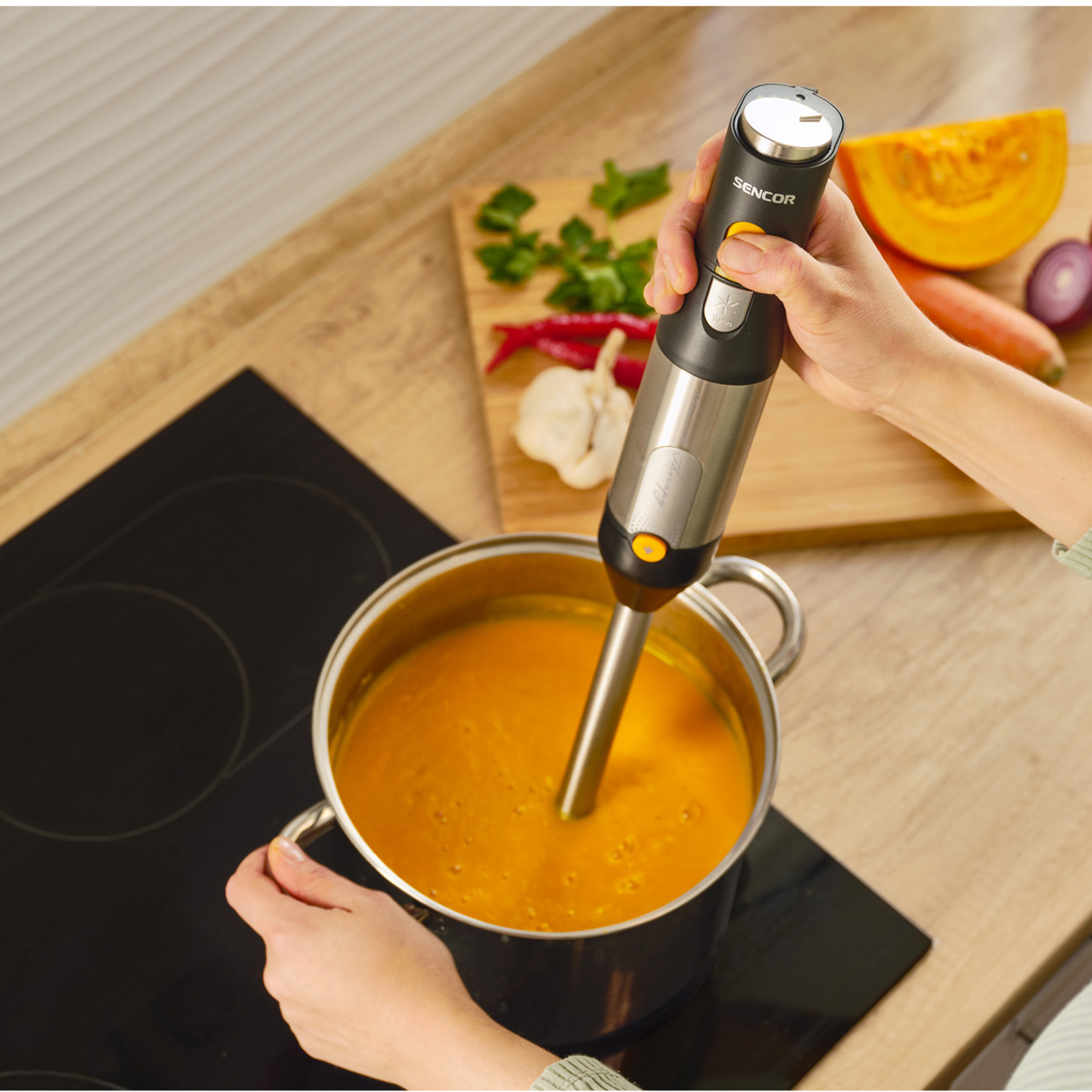 Una mano sostiene una batidora de inmersión en una olla con sopa naranja, con verduras y hierbas en la mesa.