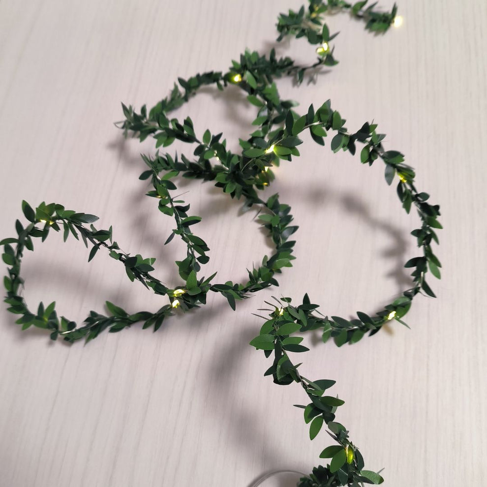 Una guirnalda de hojas artificiales verdes con pequeñas luces LED amarillas sobre una mesa de madera clara.