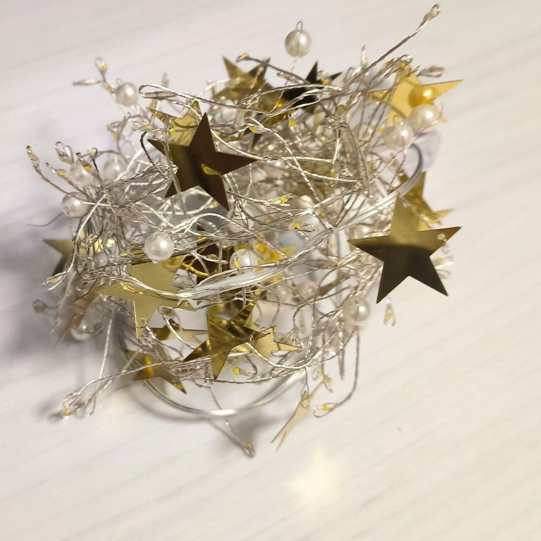Luces navideñas plateadas entrelazadas con estrellas doradas y perlas blancas sobre superficie blanca.