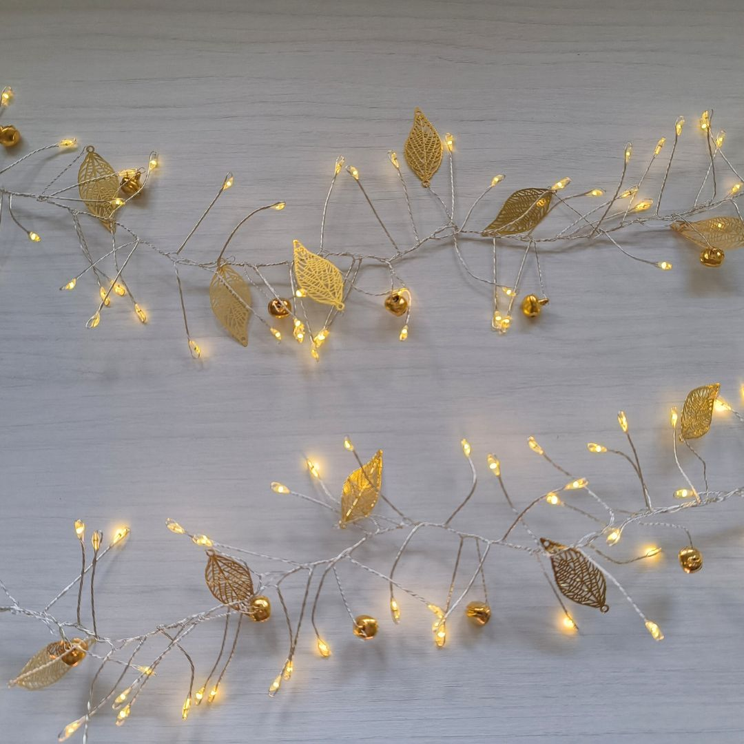 Luces de hadas con hojas doradas y pequeñas campanas sobre madera blanca.