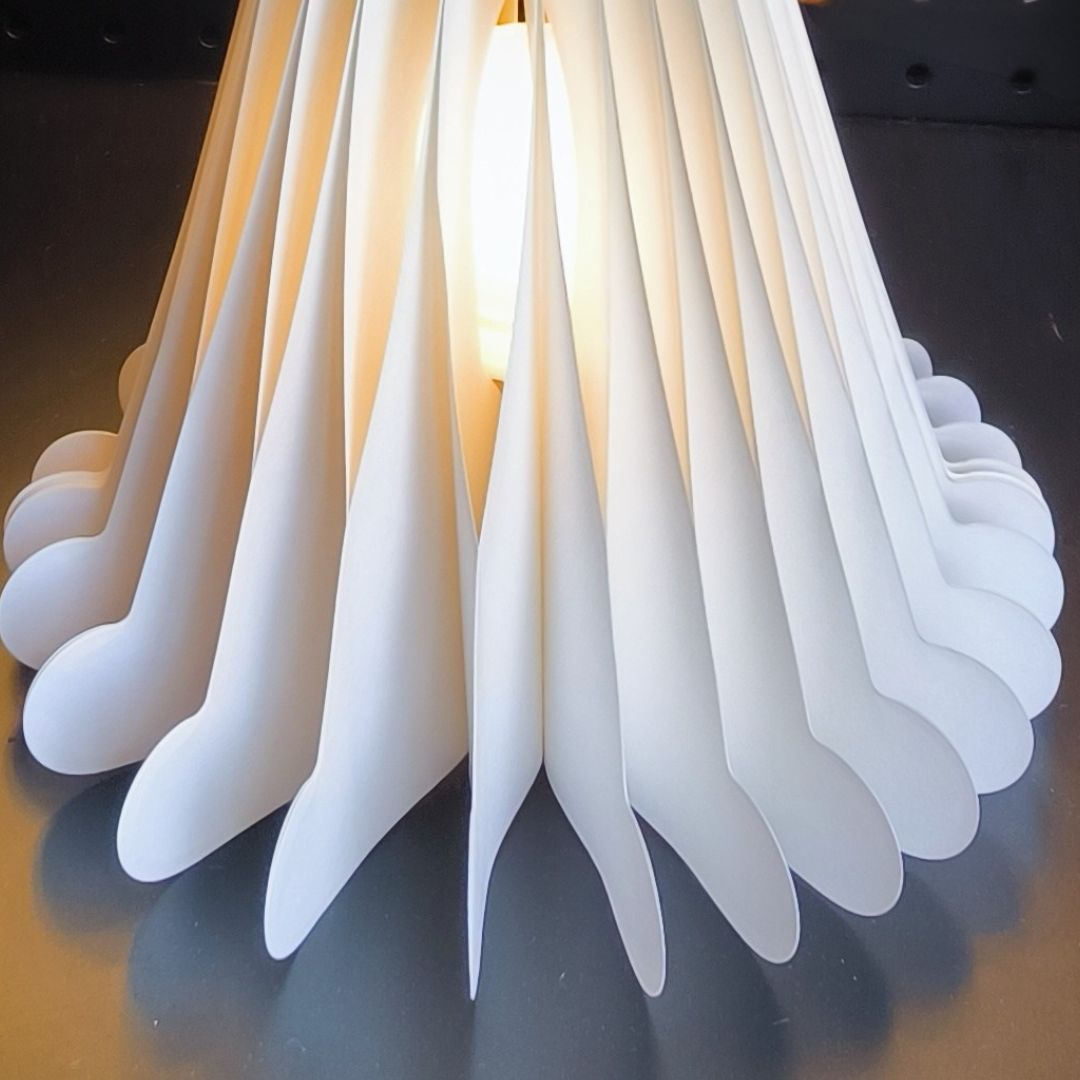 Una lámpara blanca con pantalla de hojas de papel dobladas brilla sobre una mesa oscura.