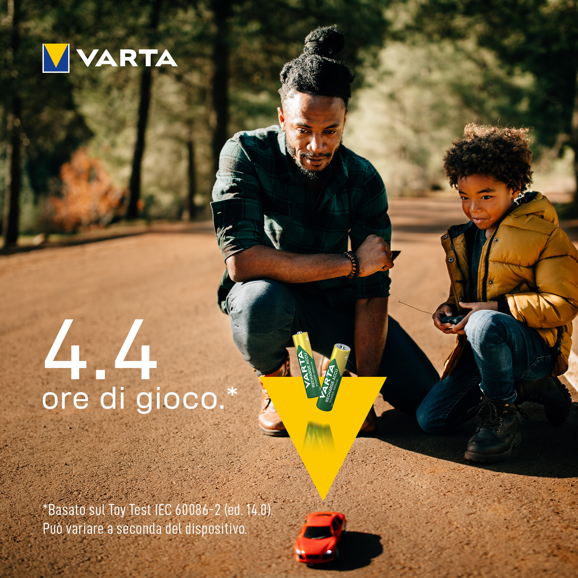 Padre e hijo se agachan en un camino de tierra, jugando con un cochecito rojo, vigilados por dos pilas Varta.