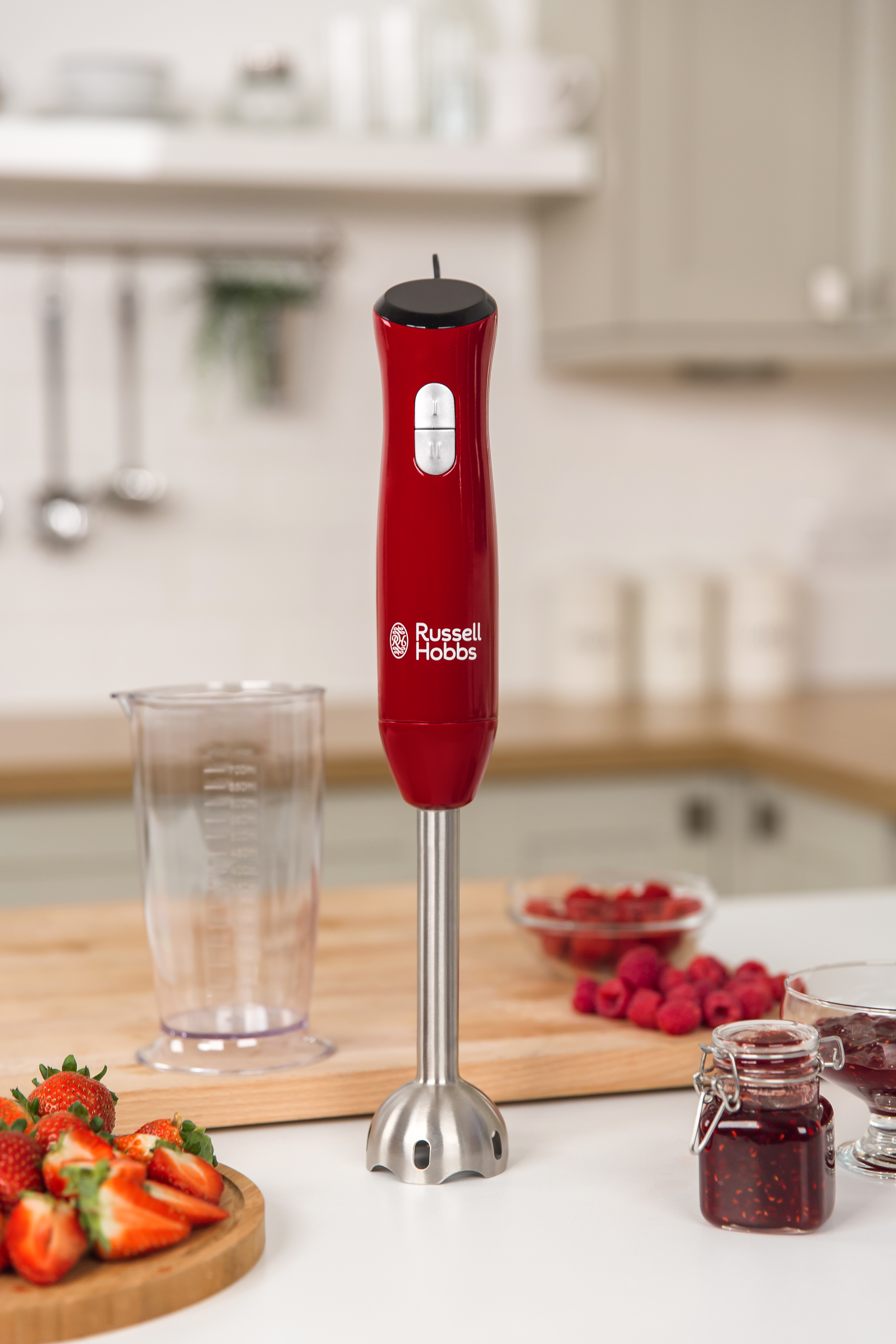Un minipimer rosso Russell Hobbs su un tagliere di legno con fragole, lamponi e vasetti di marmellata in cucina.