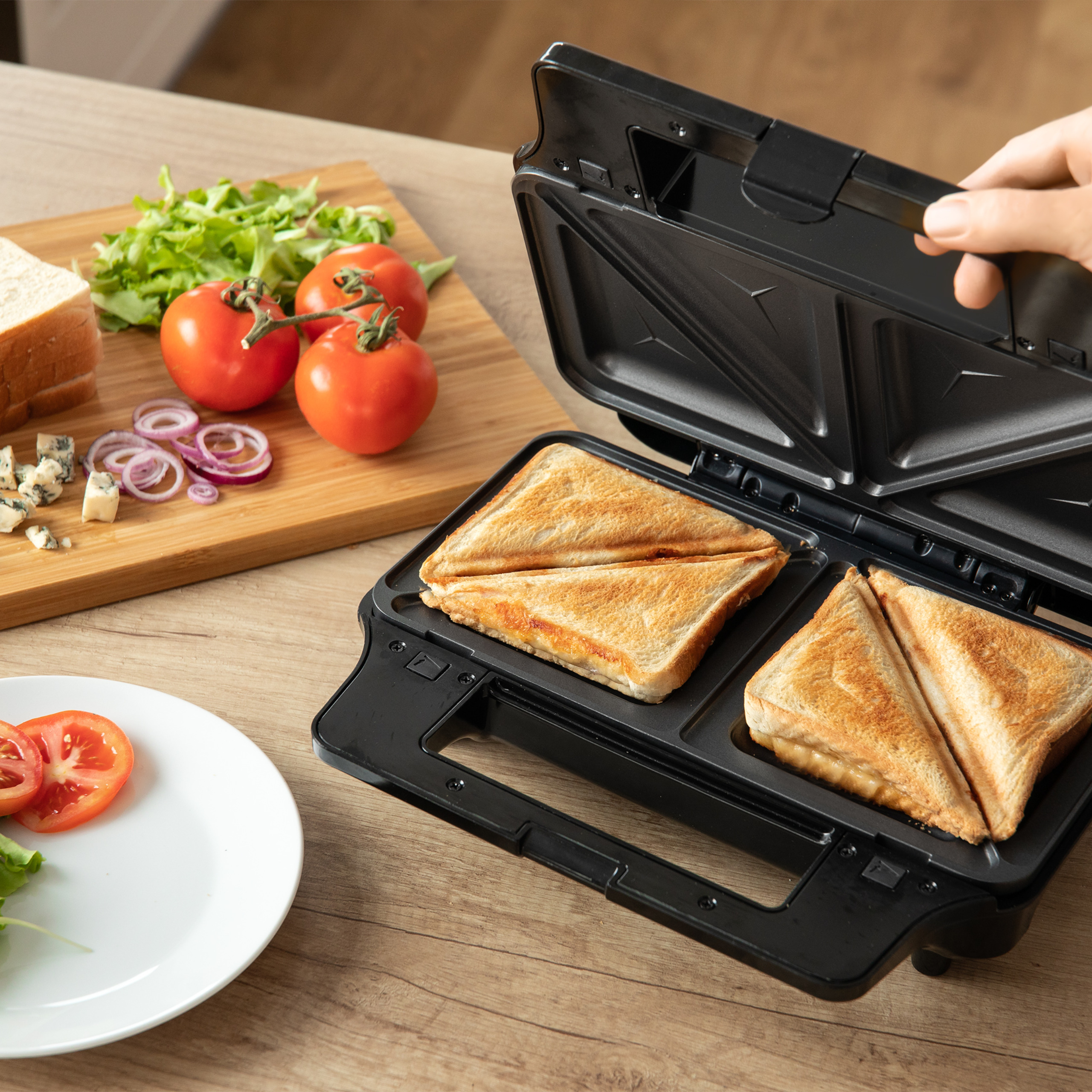 Una mano abre una sandwichera negra con sándwiches tostados dentro, junto a una tabla de cortar con verduras.