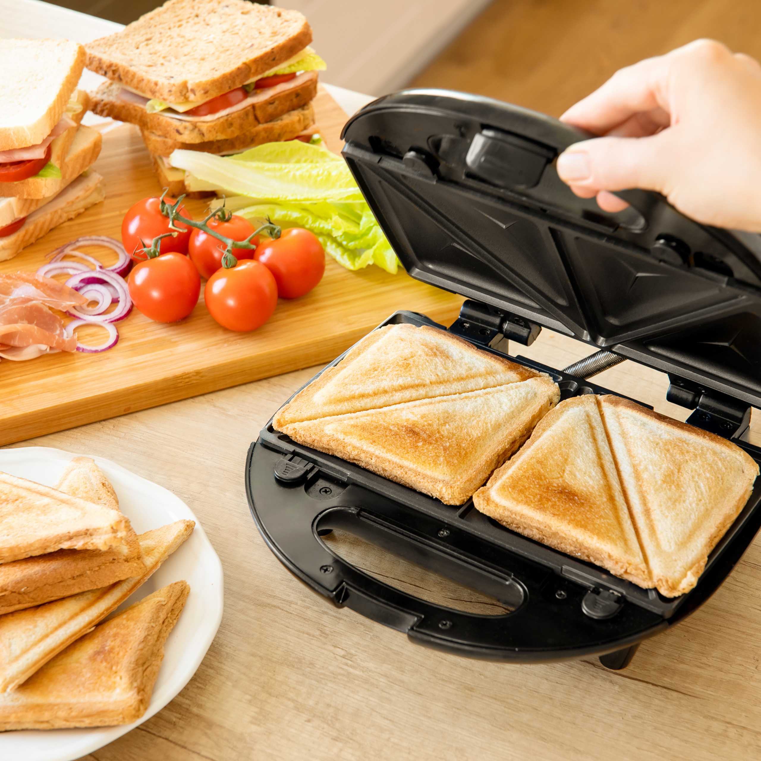Eine Hand öffnet einen schwarzen Sandwichtoaster mit getoasteten Sandwiches, neben einem Schneidebrett.