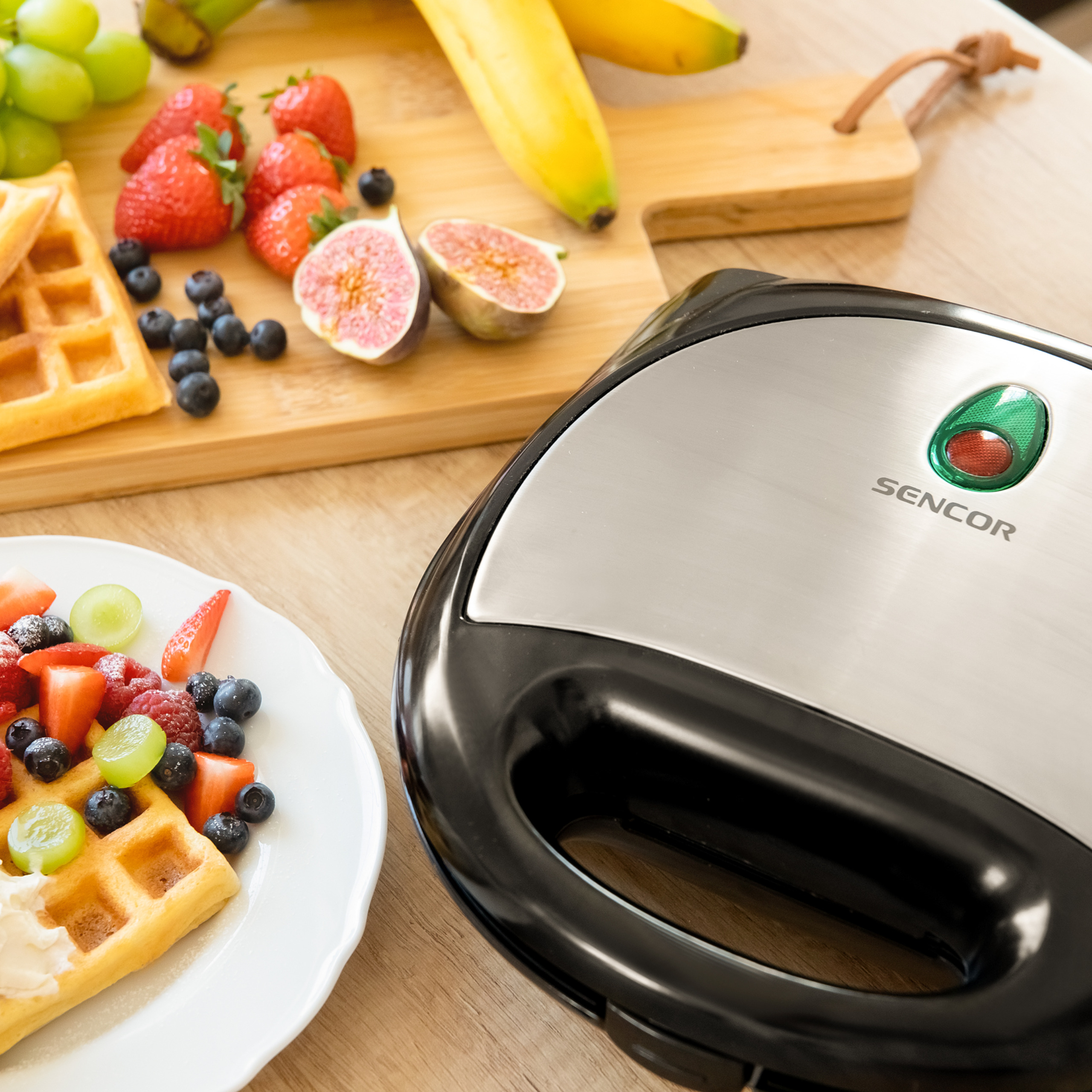 Ein Sencor-Sandwichbereiter neben einem Holzteller mit Waffeln, Früchten und Beeren auf einem Tisch.