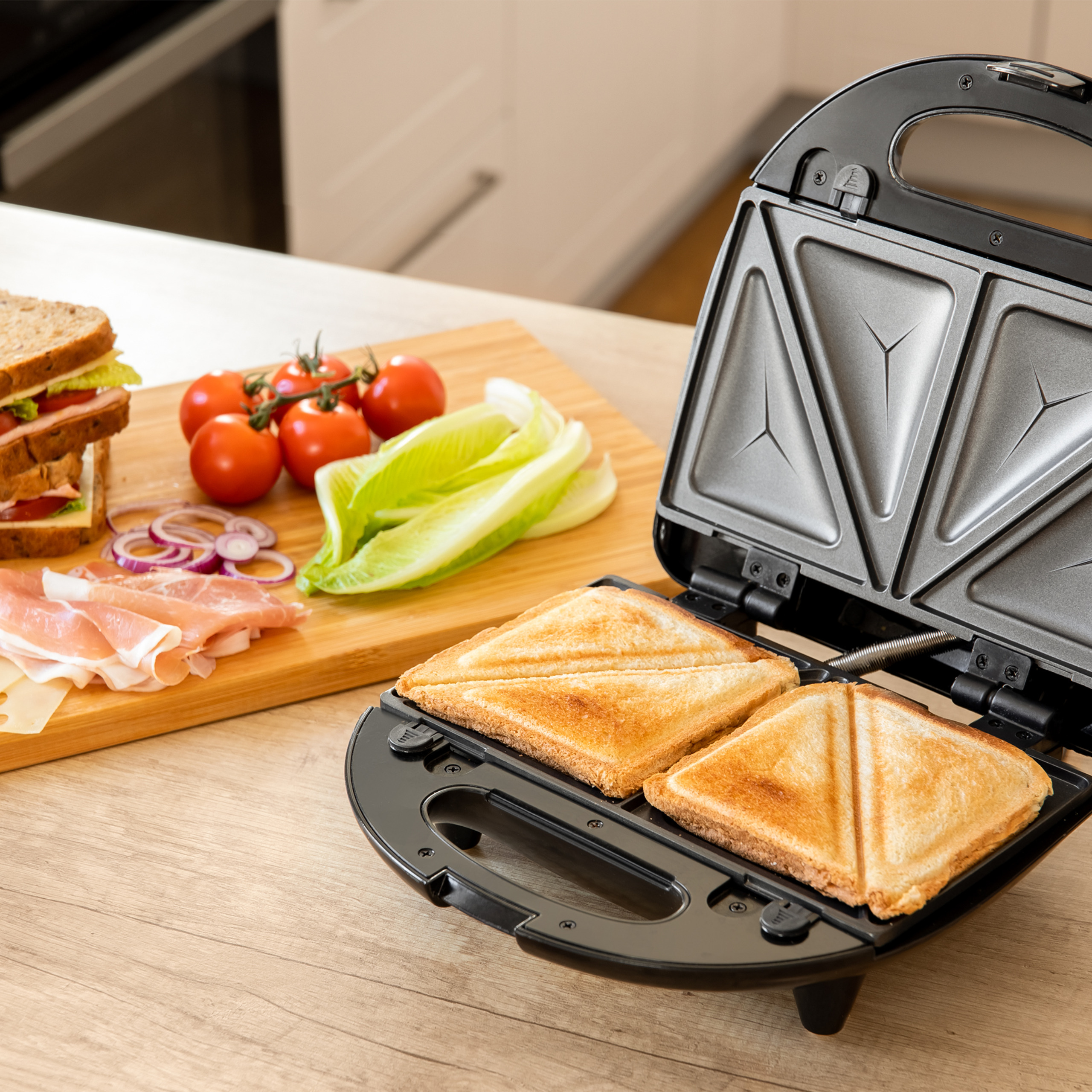 Ein offener Sandwichmaker mit zwei gegrillten Sandwiches auf einem Holztisch, daneben ein Schneidebrett mit Zutaten.
