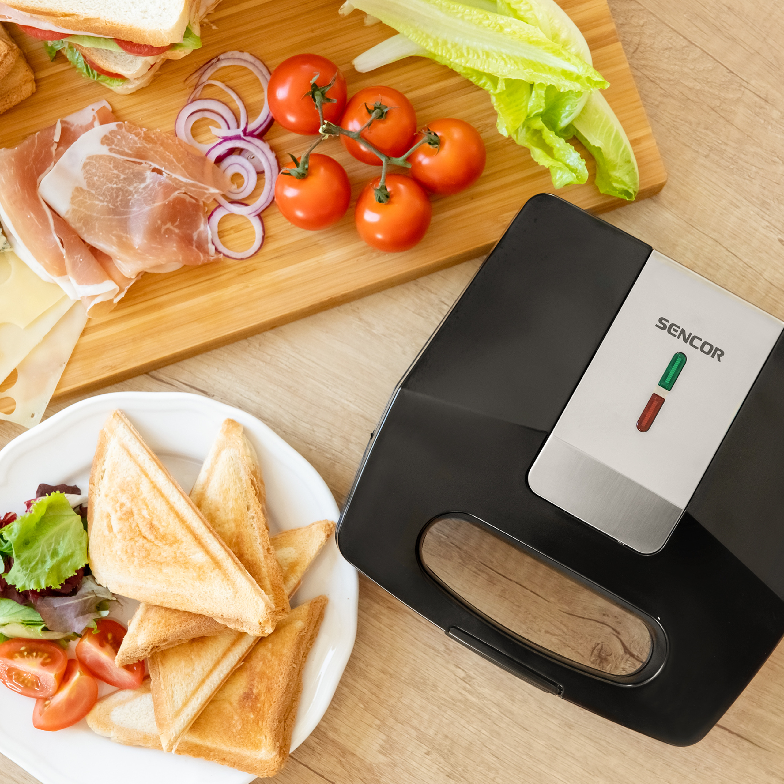 Ein Sandwichtoaster neben einem Schneidebrett mit Zutaten und einem Teller mit getoasteten Sandwiches.