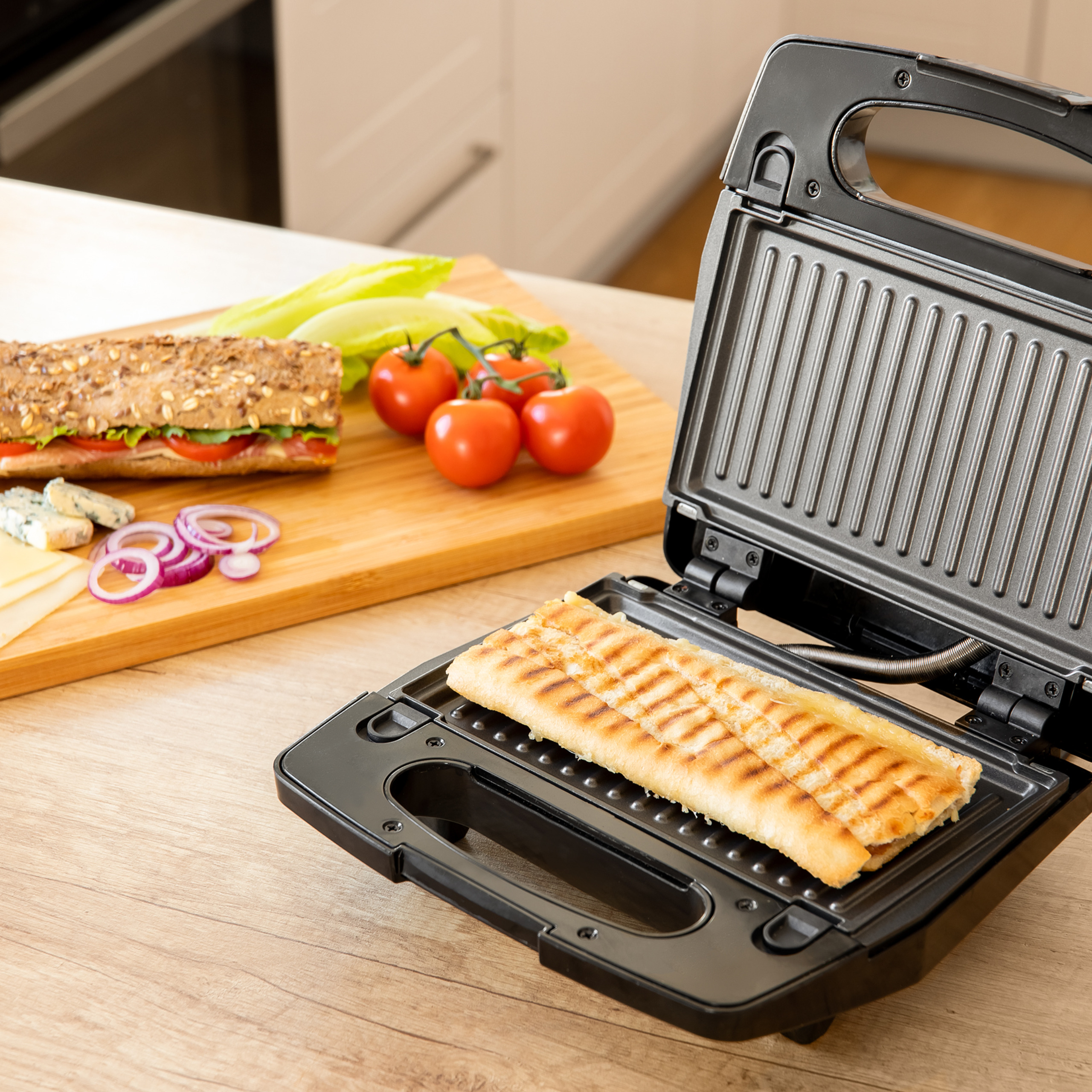 Ein Panini wird in einem offenen Kontaktgrill auf Holzoberfläche erhitzt, daneben Zutaten.