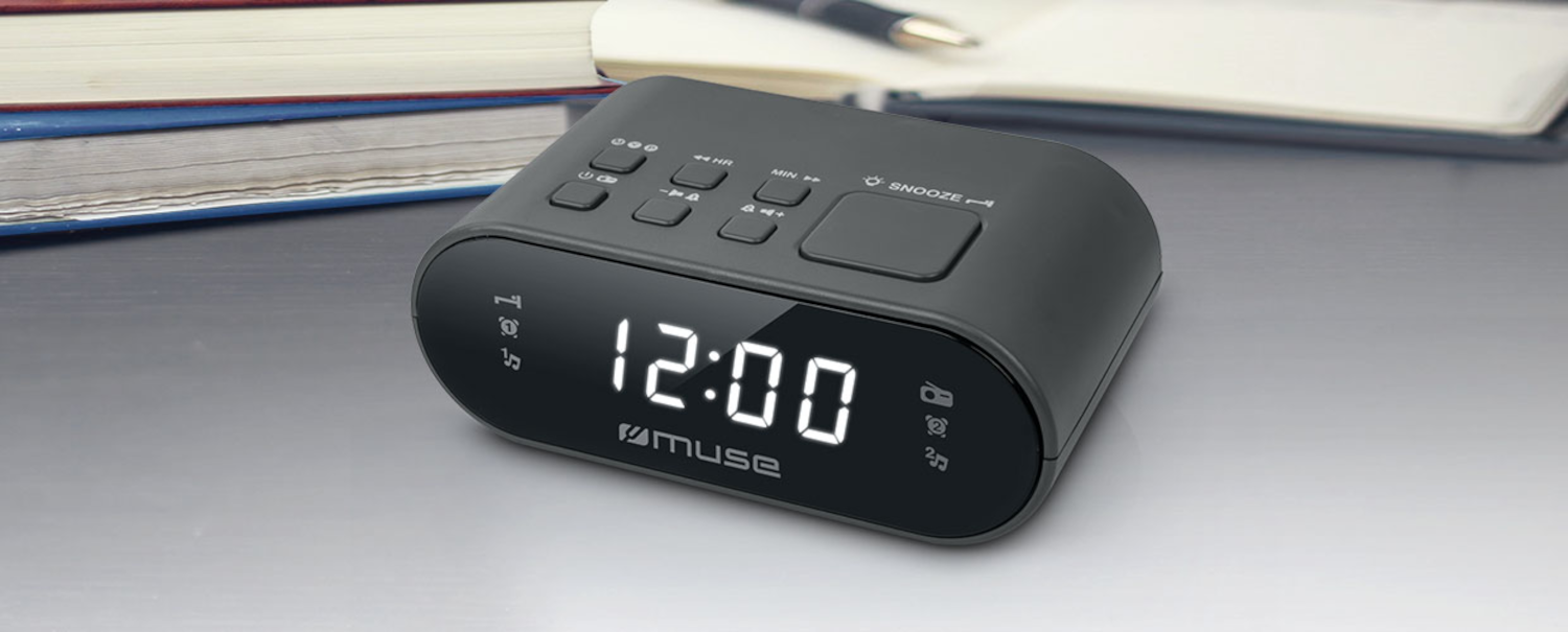 Un despertador digital Muse negro que muestra las 12:00 en un escritorio junto a libros y un bolígrafo.