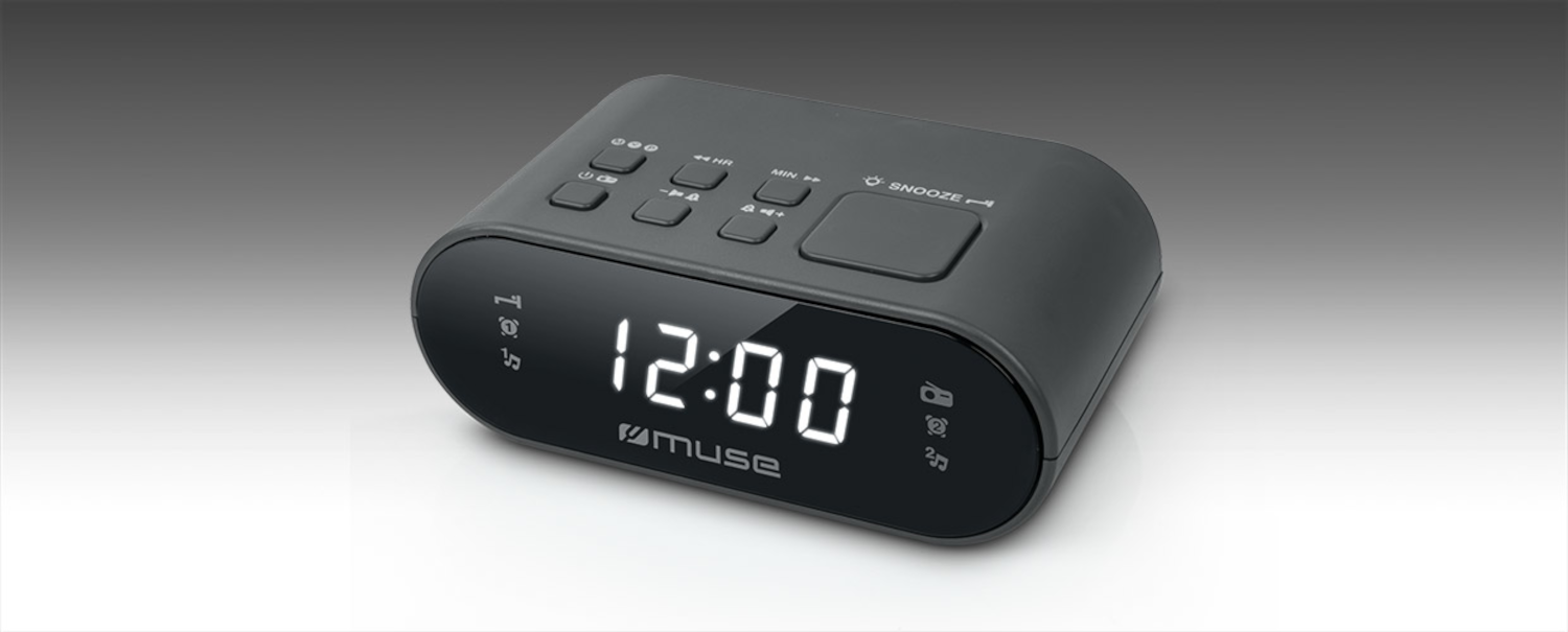 Un despertador digital negro con dígitos blancos brillantes que muestran las 12:00, botones de control, fondo gris.