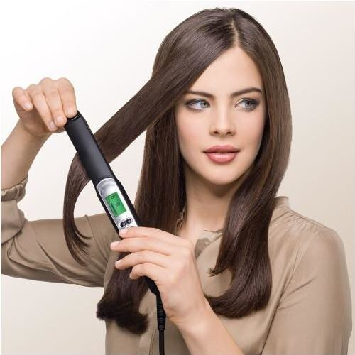 Una donna con lunghi capelli castani liscia una ciocca di capelli usando una piastra nera con display verde.