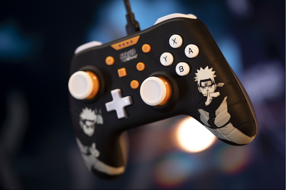 Un mando de juego negro de Naruto con botones blancos y naranjas y dos joysticks, sobre un fondo oscuro.