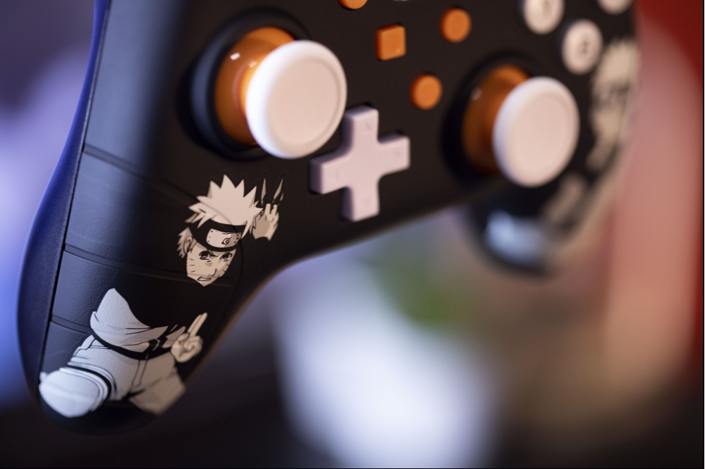 Un controlador de juegos negro con un adhesivo de Naruto, joysticks blancos y detalles naranjas.