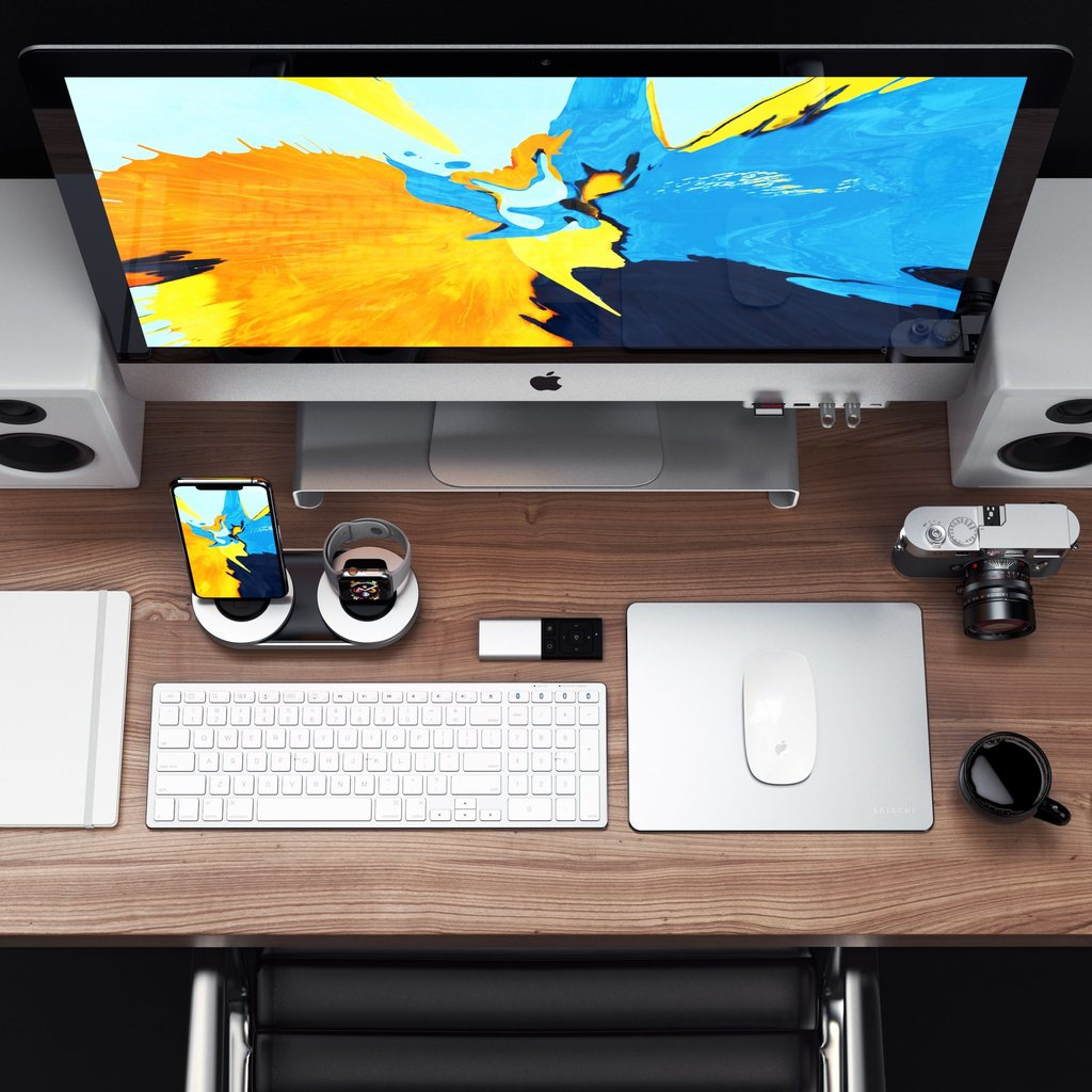 Un escritorio de madera con un iMac, teclado blanco, ratón, cargador dual, cámara antigua, libreta y taza de café.