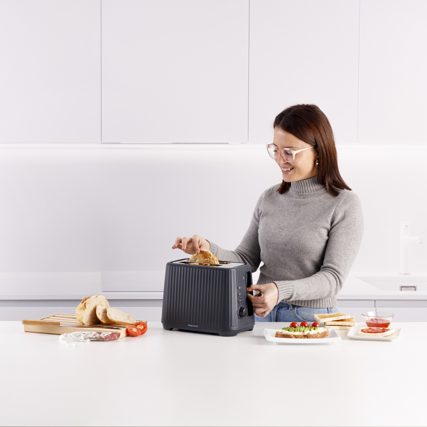 Kobieta w szarym swetrze wyjmuje tost z tostera w białej kuchni.
