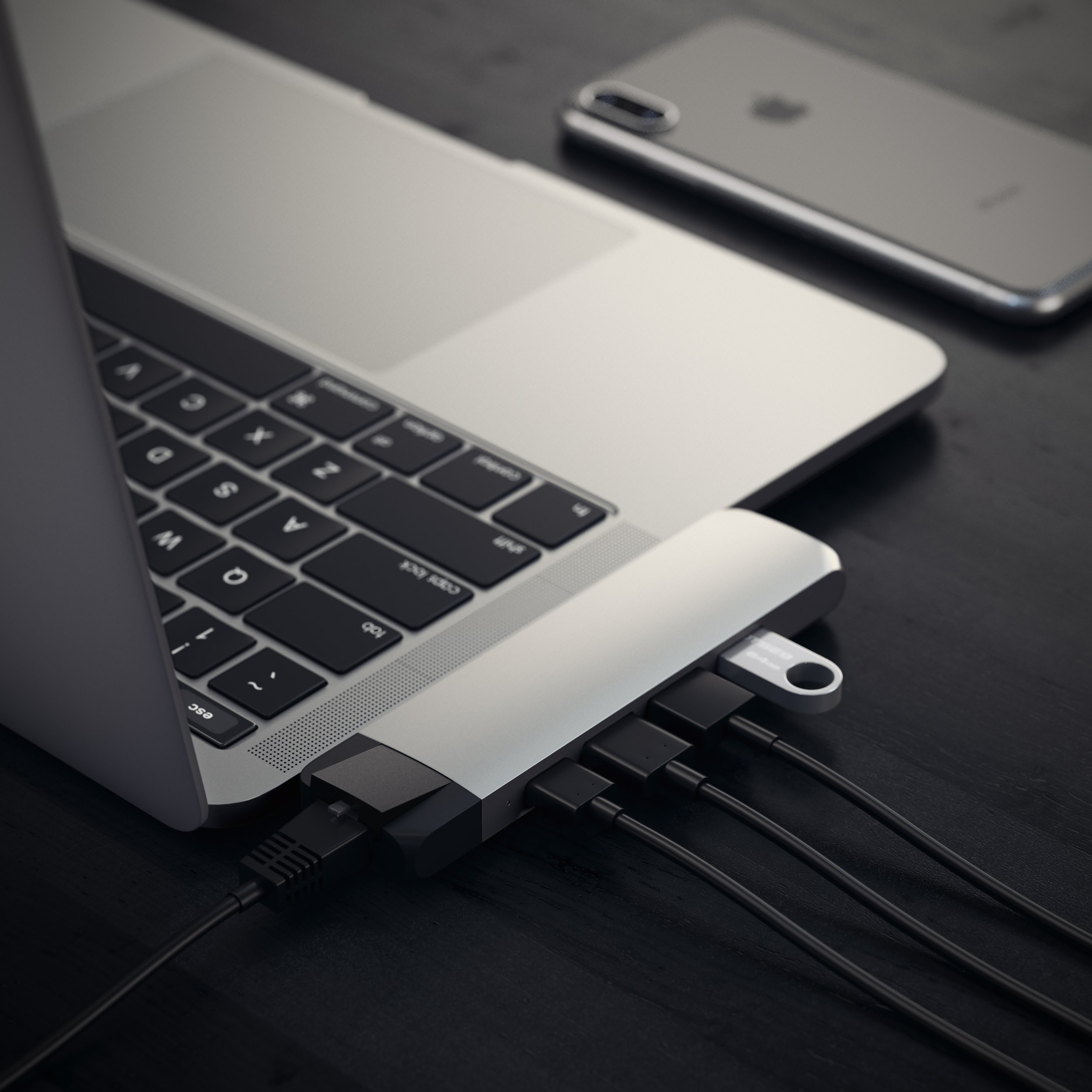 A silver laptop with a connected USB-C hub and cables on a dark table, with a smartphone nearby.