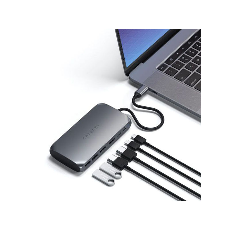 Concentrador USB Satechi gris con cables conectados y unidades USB junto a un portátil sobre fondo blanco.
