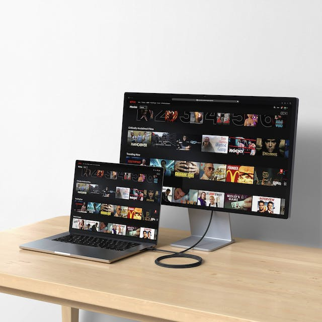 Un ordenador portátil y un monitor con una plataforma de streaming de películas están en un escritorio de madera.