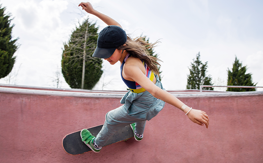 Eine junge Skaterin in schwarzer Kappe und grauem Overall zeigt einen Trick in einer rosafarbenen Skatebowl.