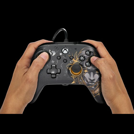 Hände halten einen schwarzen kabelgebundenen Xbox-Controller mit gold-grauer Grafik auf schwarzem Grund.