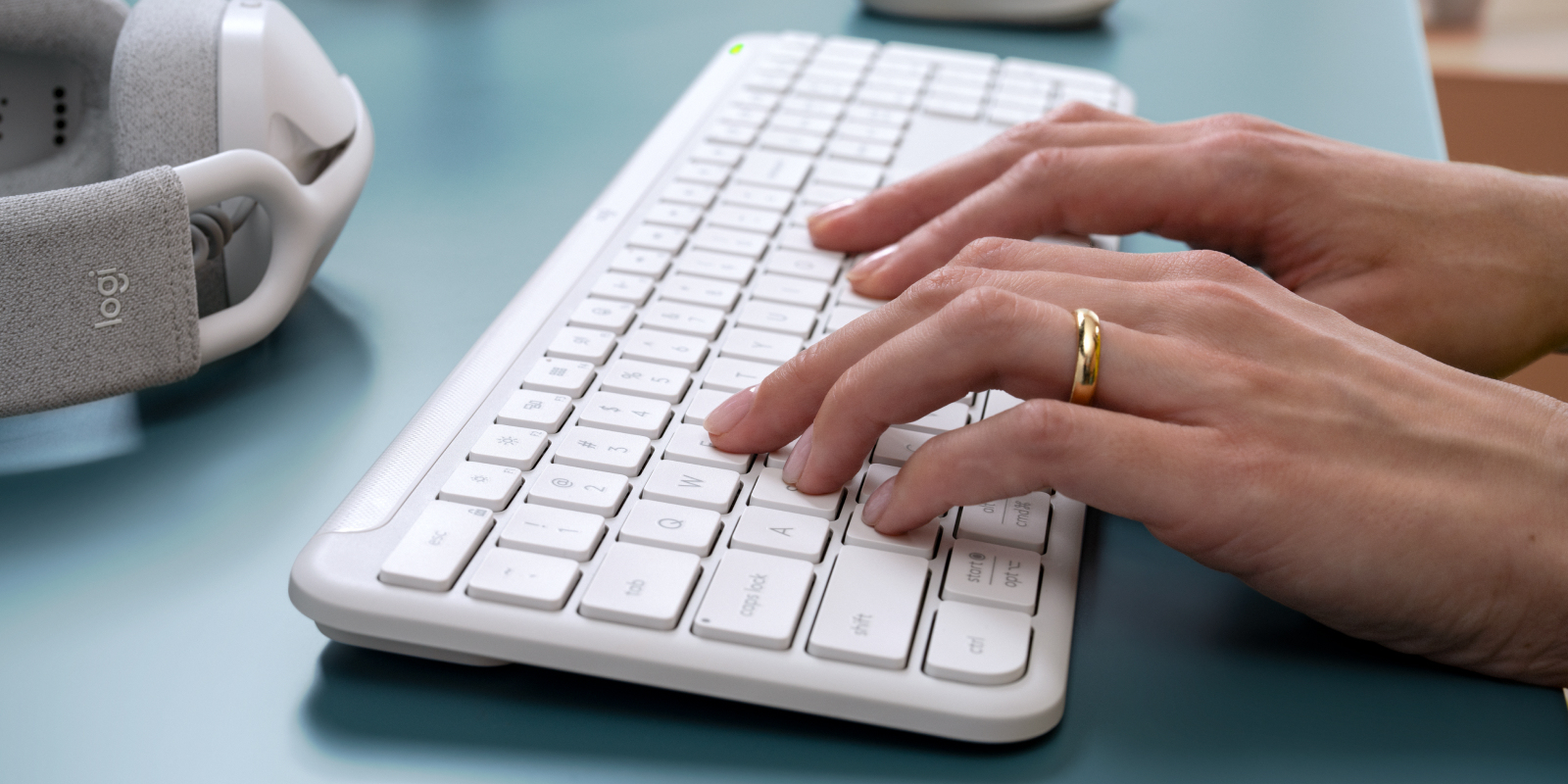 Persona escribiendo en un teclado blanco en un escritorio azul con auriculares a la izquierda.