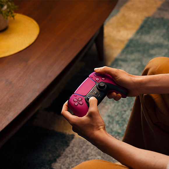 Eine Person sitzt und hält einen pinken PlayStation-Controller in den Händen, neben einem Holztisch.
