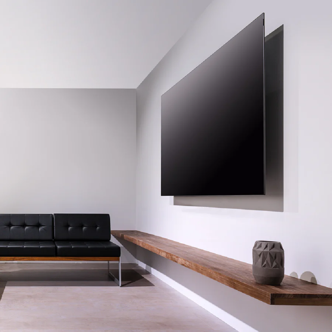 Un televisor negro grande cuelga en una pared blanca sobre un estante de madera largo con un jarrón gris.