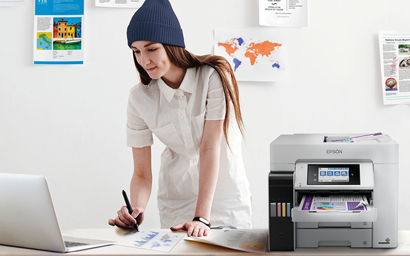 Eine Frau in weißer Bluse und blauer Mütze schreibt am Tisch vor einem Laptop. Neben ihr steht ein Epson-Drucker.