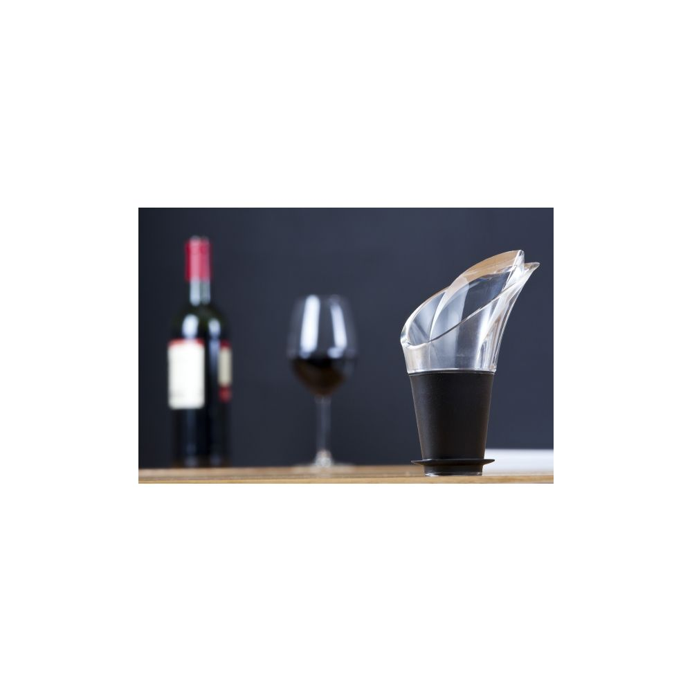Un areatore per vino in vetro con base nera su un tavolo di legno; sullo sfondo una bottiglia e un calice.