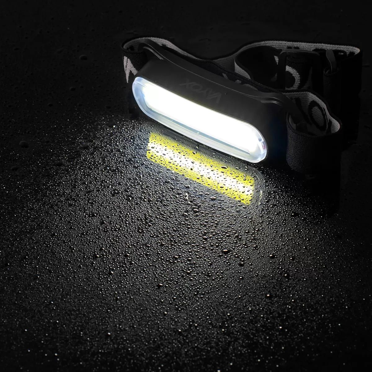 Egy bekapcsolt LED-es fejlámpa pihen egy vízcseppekkel borított, nedves, sötét felületen.