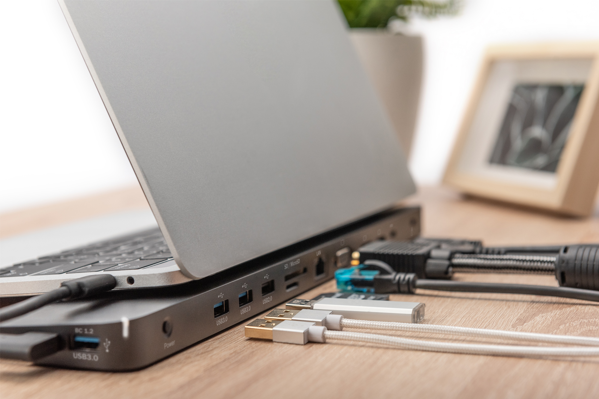 Ein silberner Laptop an einer Dockingstation mit mehreren Kabeln und USB-Sticks auf Holzschreibtisch.