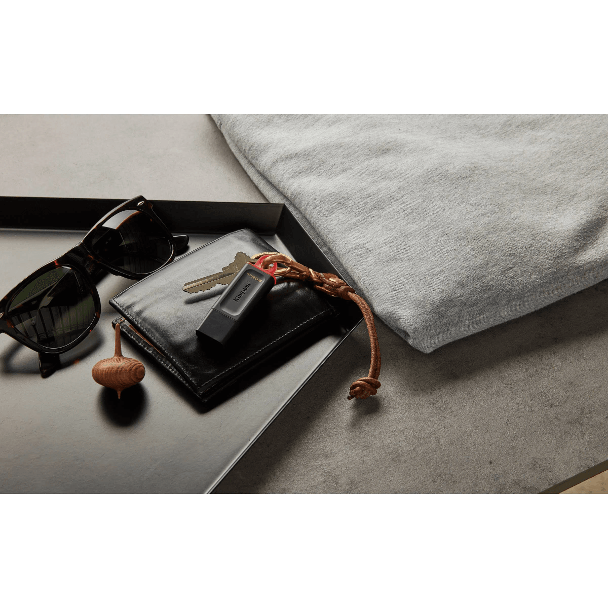 Gafas de sol, cartera, llaves, memoria USB y una peonza de madera en una bandeja junto a un suéter gris.