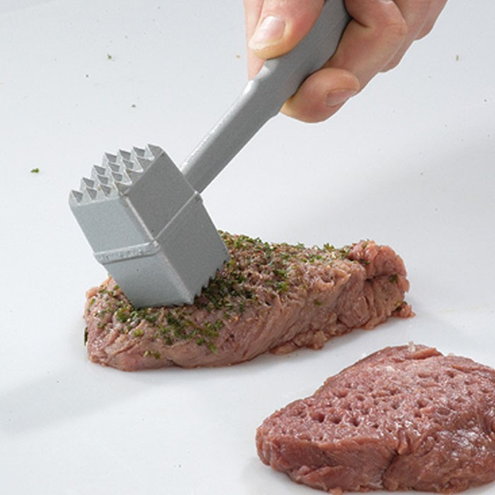 Una mano usa un batticarne su una bistecca condita con erbe su una superficie bianca.