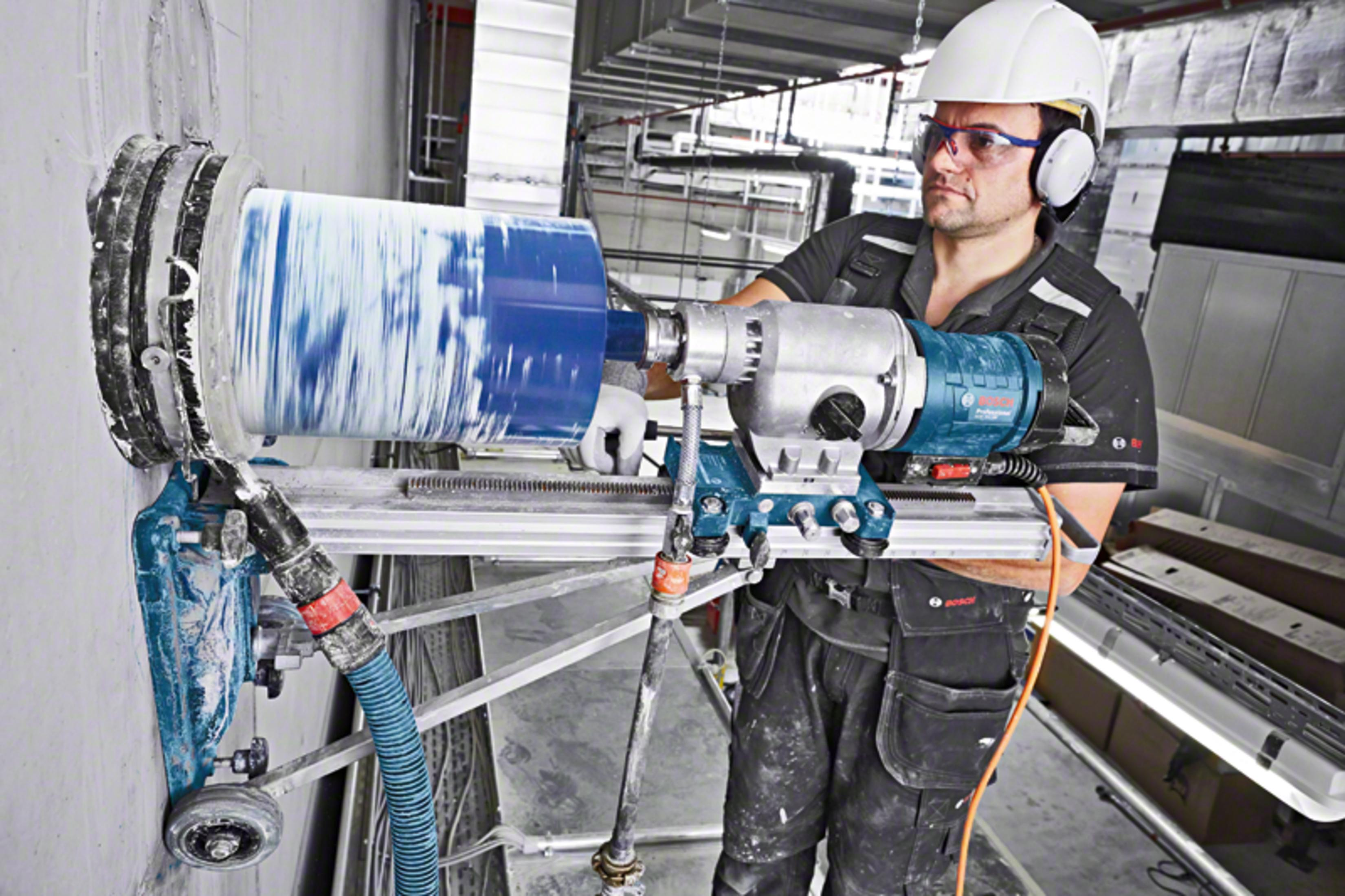Un trabajador con casco blanco y gafas de seguridad opera una gran máquina perforadora en una pared.
