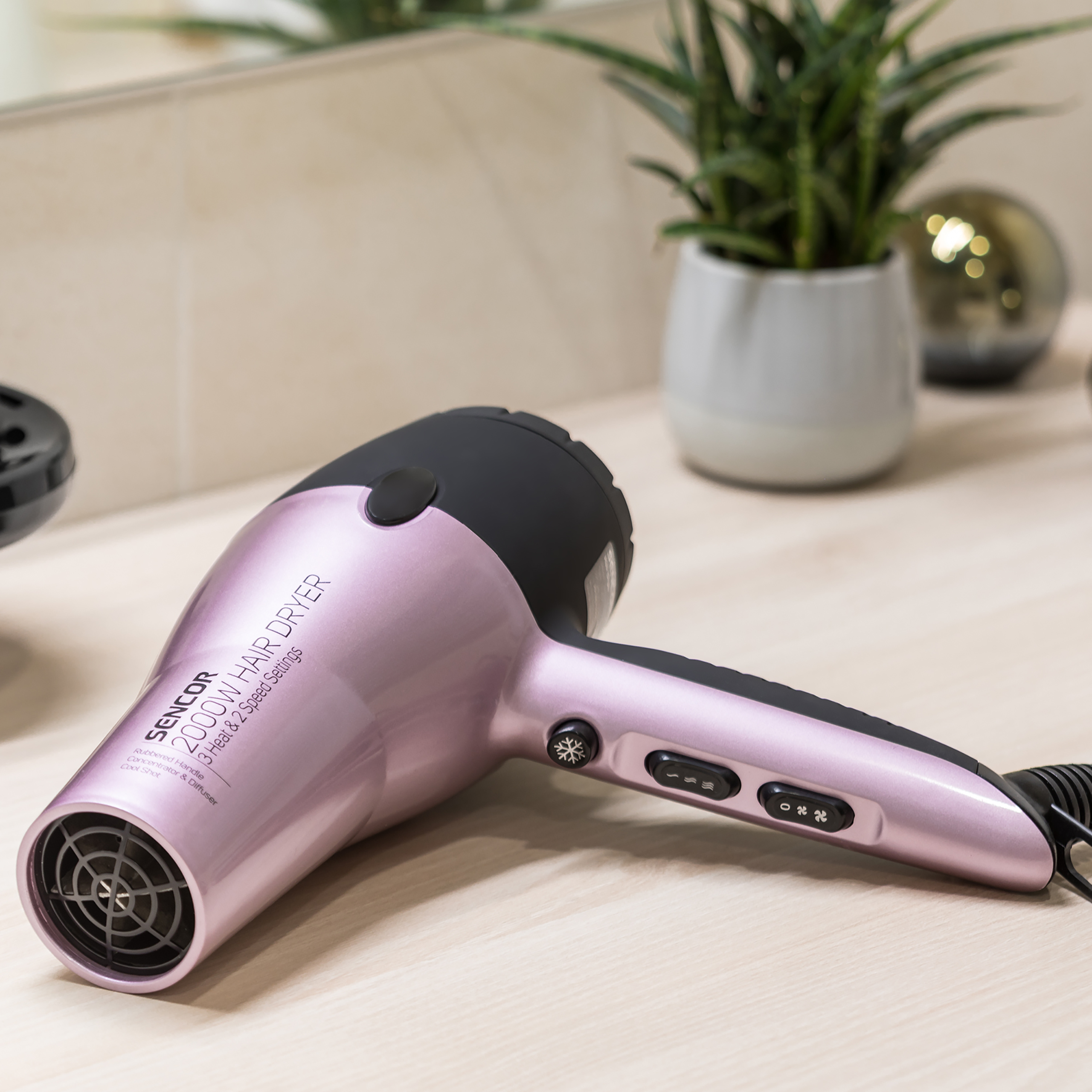 A pink and black hair dryer on a light wooden table, with a potted plant and mirror in the blurred background.