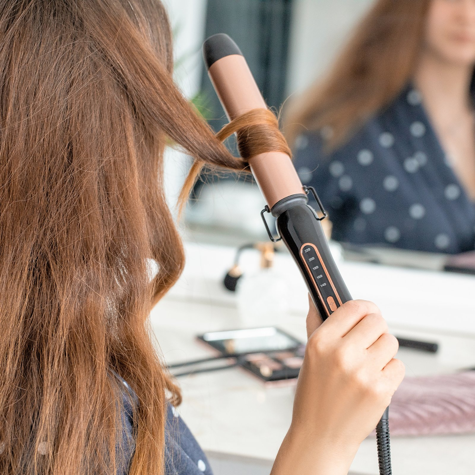 Eine Frau lockt ihre braunen Haare mit einem schwarzen Lockenstab vor einem Spiegel.