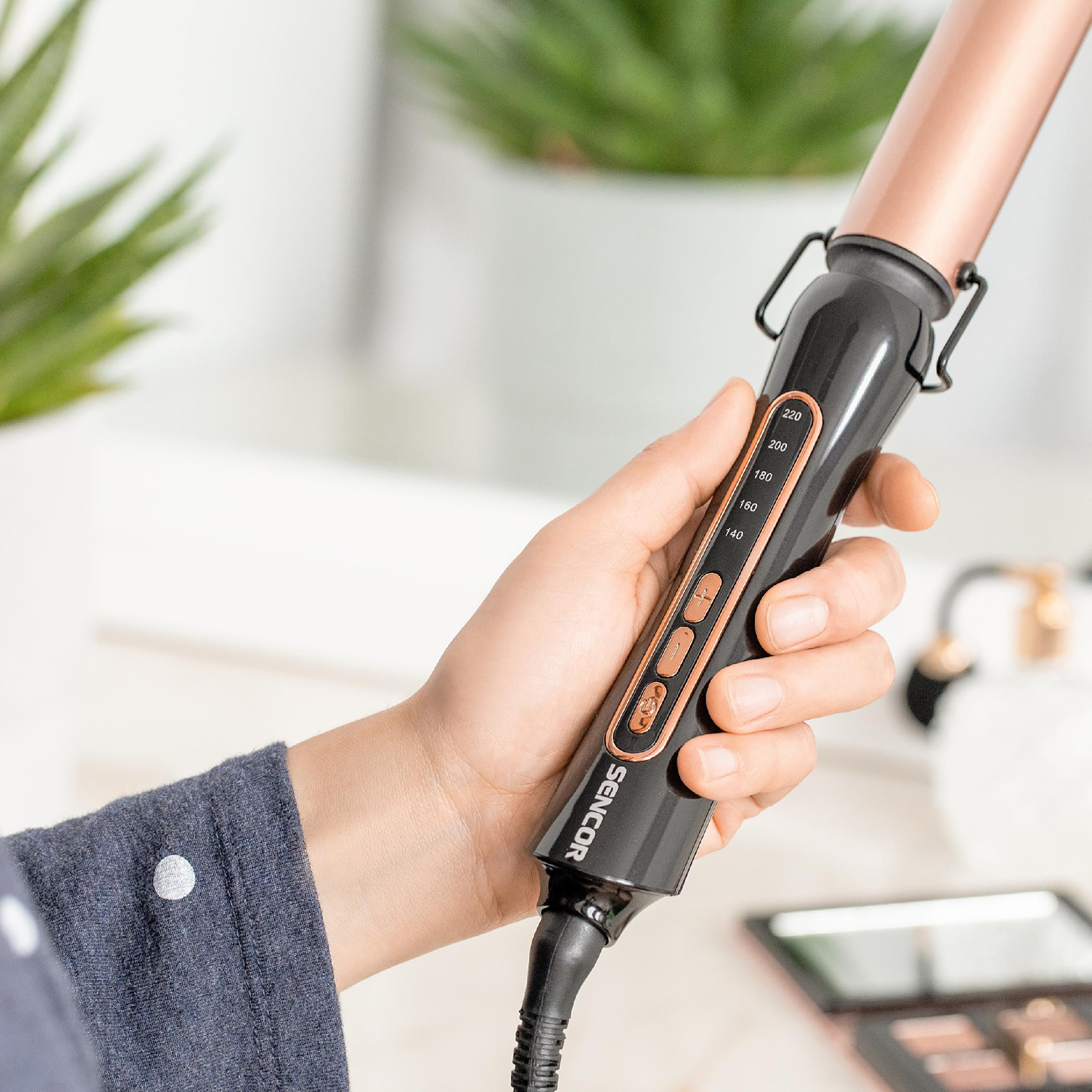 Eine Hand hält einen schwarz-roségoldenen Lockenstab mit digitaler Temperatureinstellung vor verschwommenem Hintergrund.