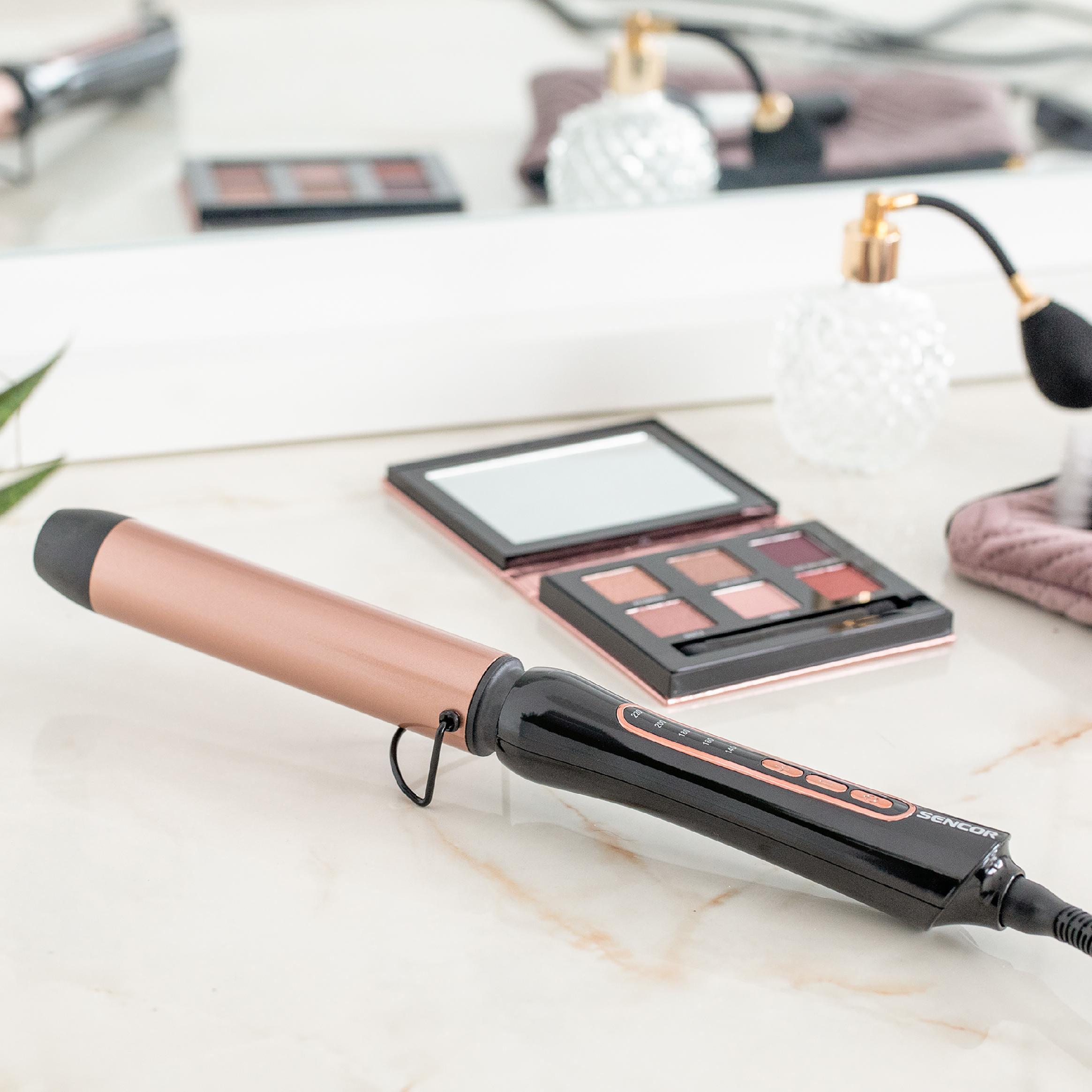 Ein roségoldener und schwarzer Lockenstab liegt auf einem Marmortisch, mit Make-up-Paletten im Hintergrund.