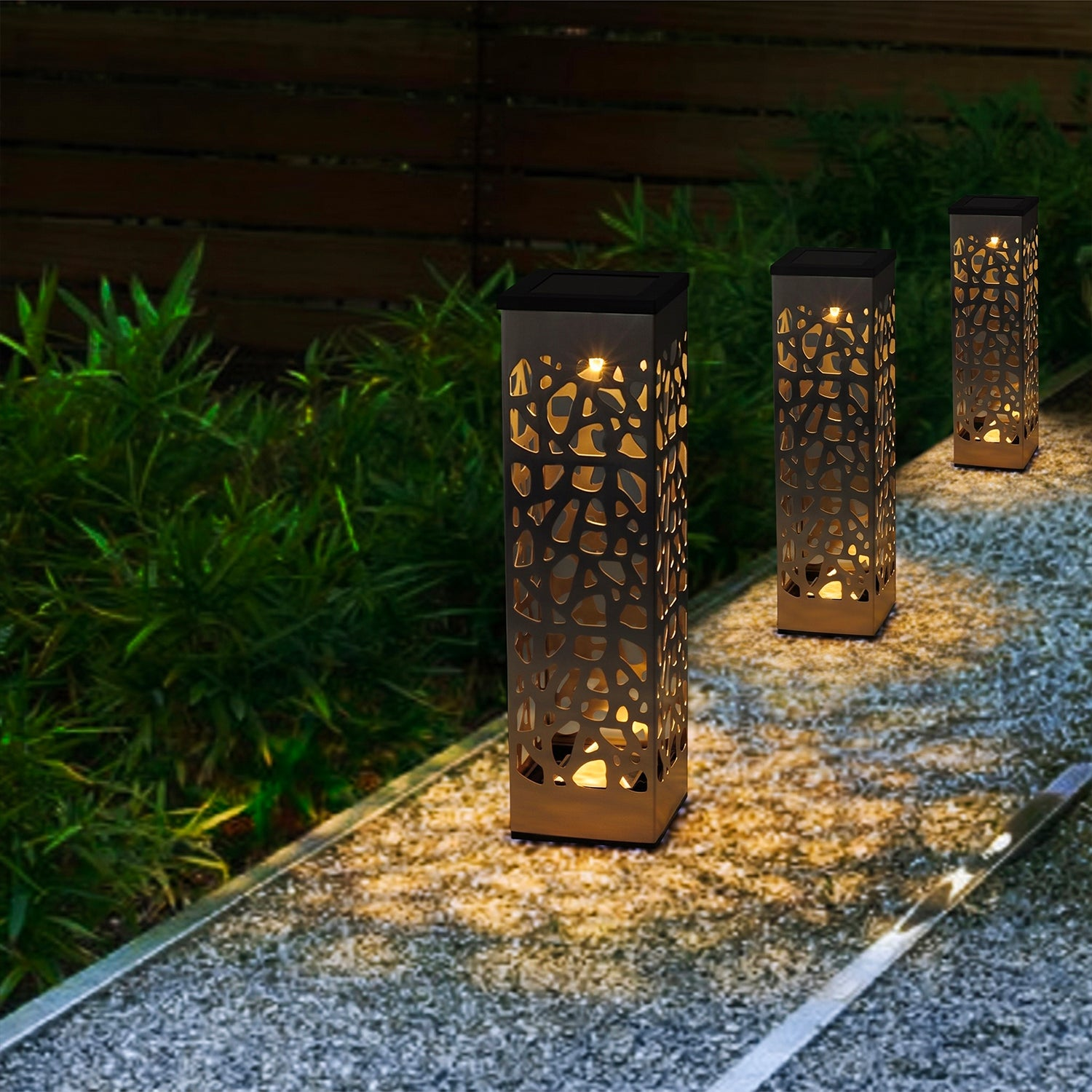 Tre luci a pilastro illuminate a motivi si trovano su un sentiero di ghiaia in un giardino notturno con piante.