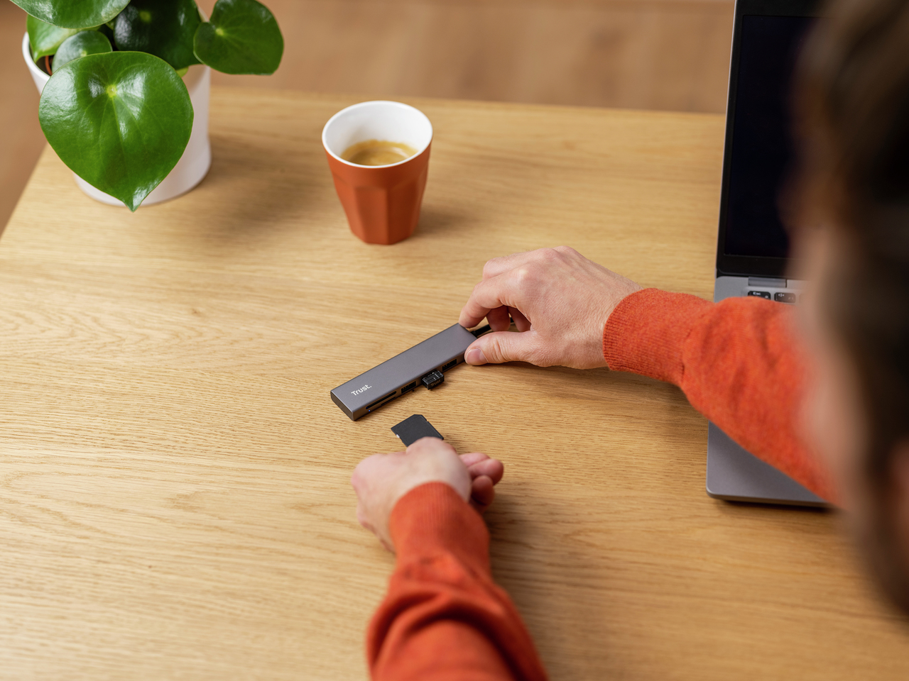 Person in red sweater connecting a hub and SD card on wooden desk with laptop.