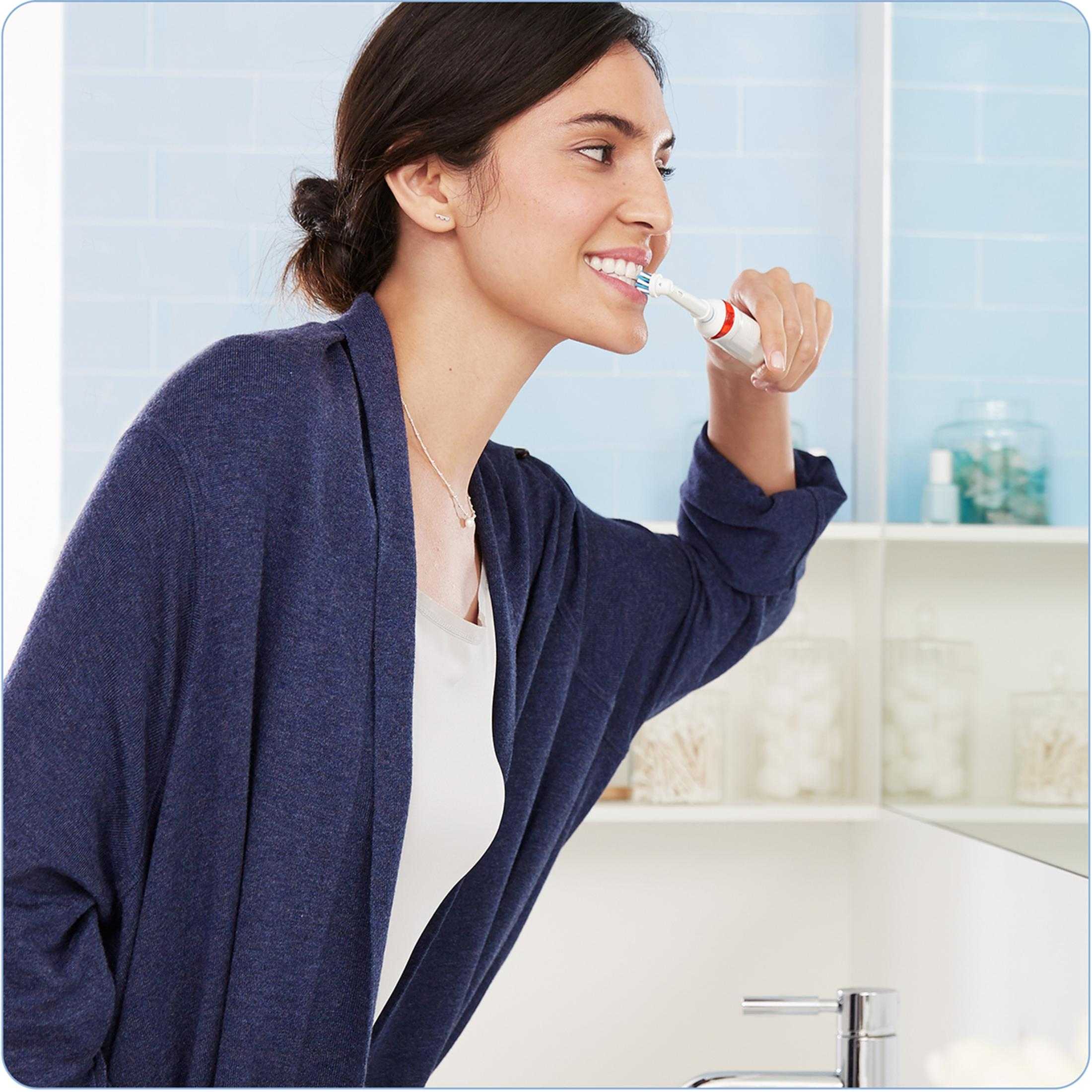 Una donna sorridente con un cardigan blu si lava i denti con uno spazzolino elettrico davanti allo specchio del bagno.