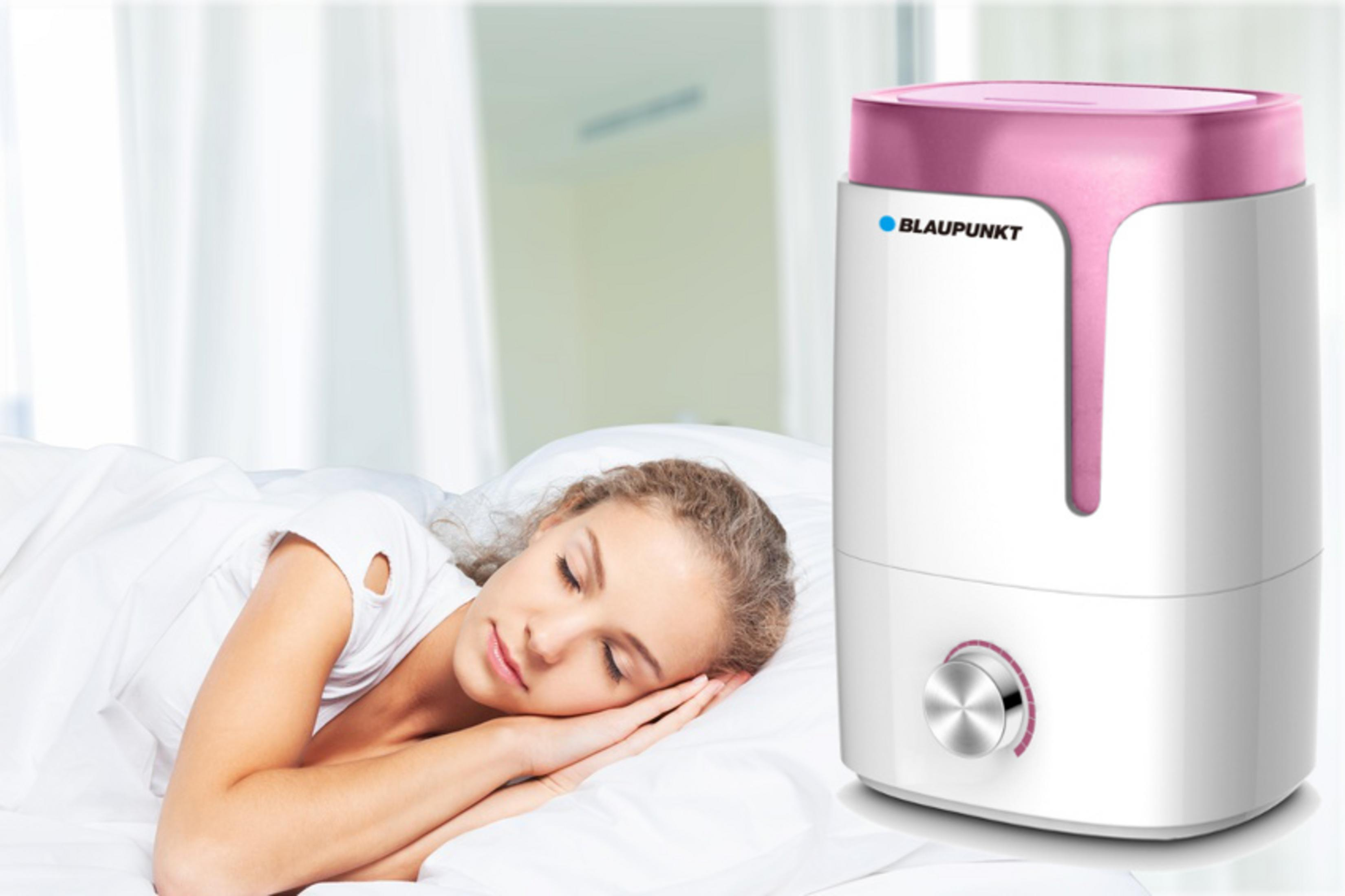 Un humidificador Blaupunkt blanco y rosa sobre una mesa mientras una mujer joven duerme plácidamente al fondo.