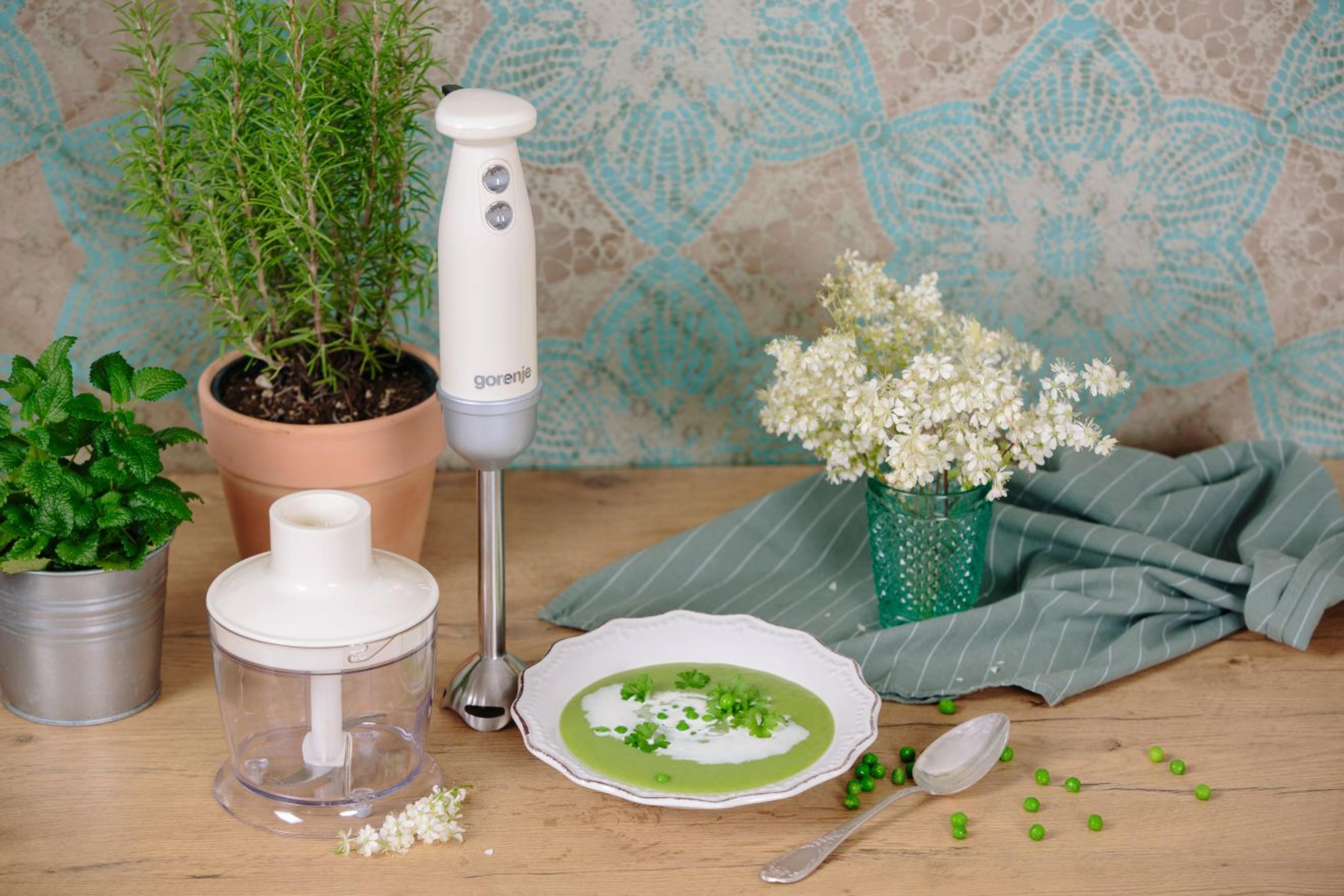 Ein weißer Stabmixer steht auf einem Holztisch neben einer Schüssel grüner Suppe, Kräutern und Blumen.