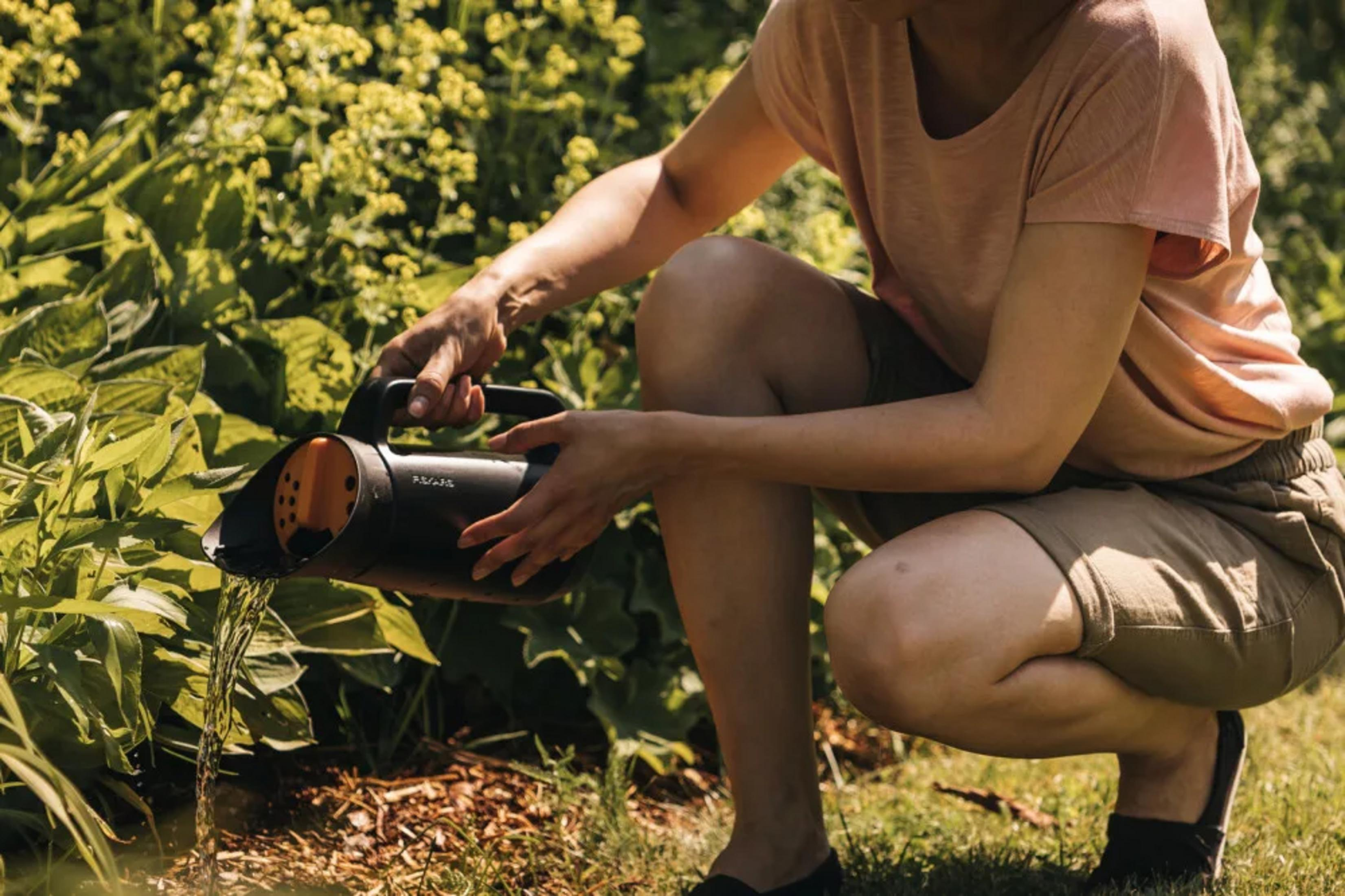 Eine Frau kniet im Garten und gießt Pflanzen mit einer schwarzen Gießkanne.