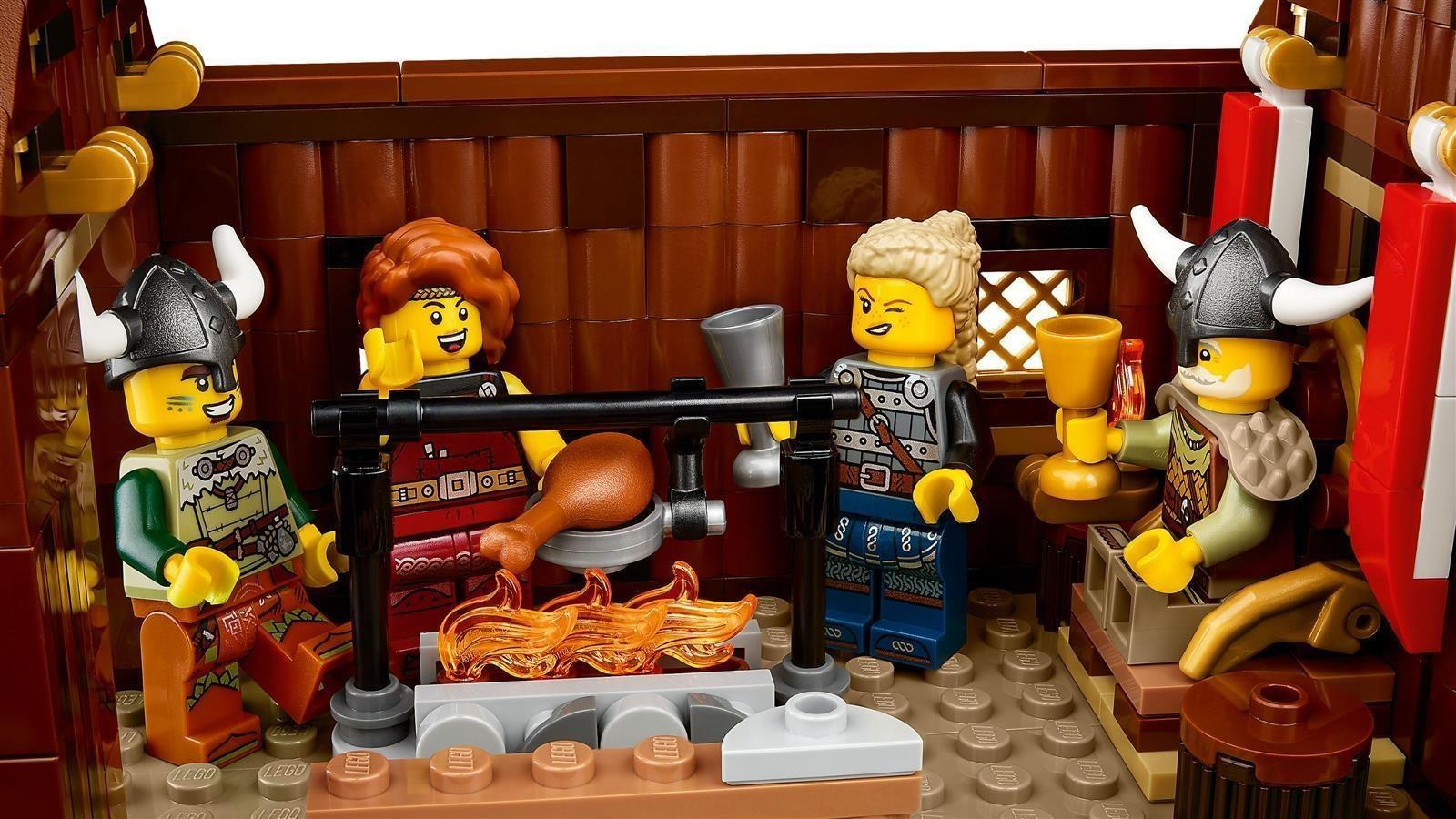 Vier Lego-Wikingerfiguren sitzen um eine Feuerstelle in einer Holzhütte und feiern mit einem Hähnchenschenkel.