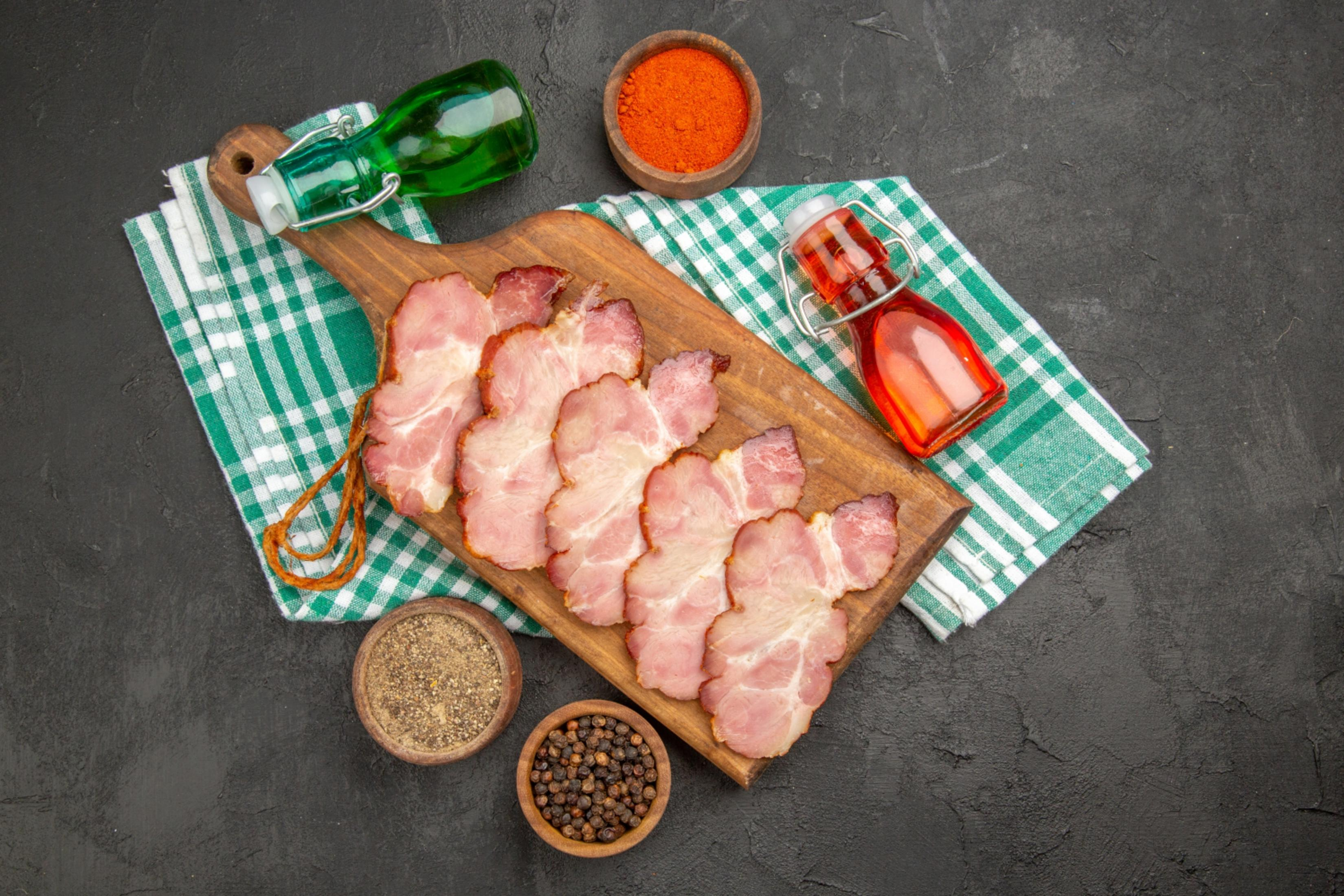 Fette di prosciutto su tagliere di legno con spezie e bottiglie d'olio su tovaglia a quadretti verde e fondo scuro.