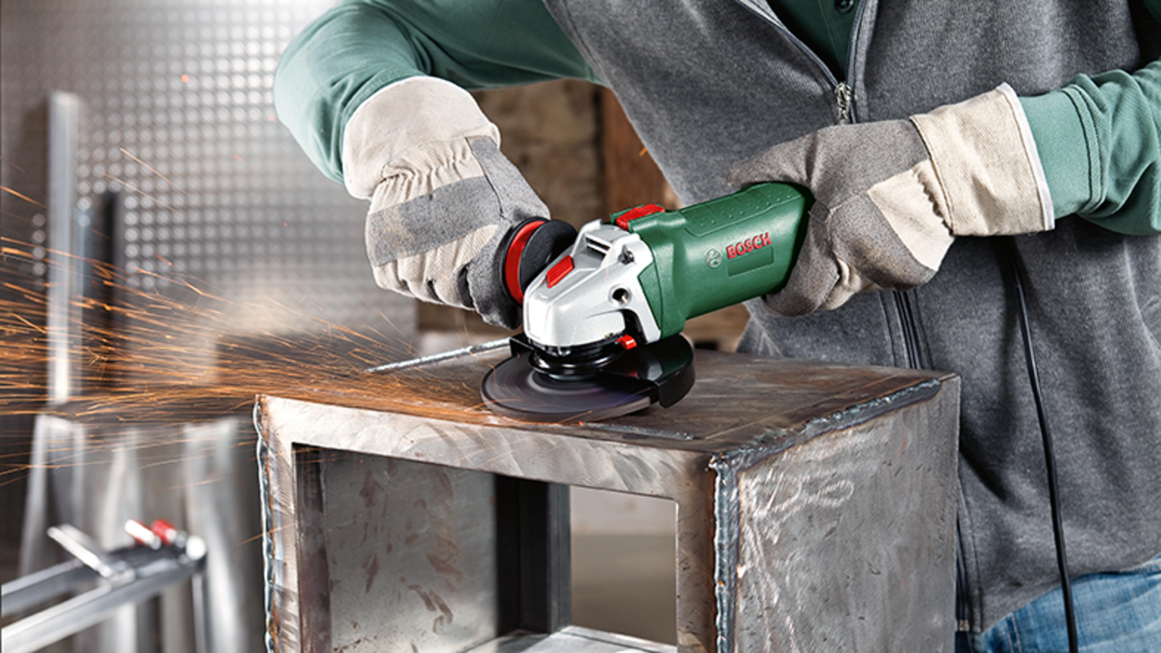 Un trabajador con guantes protectores usa una amoladora angular verde, creando chispas.
